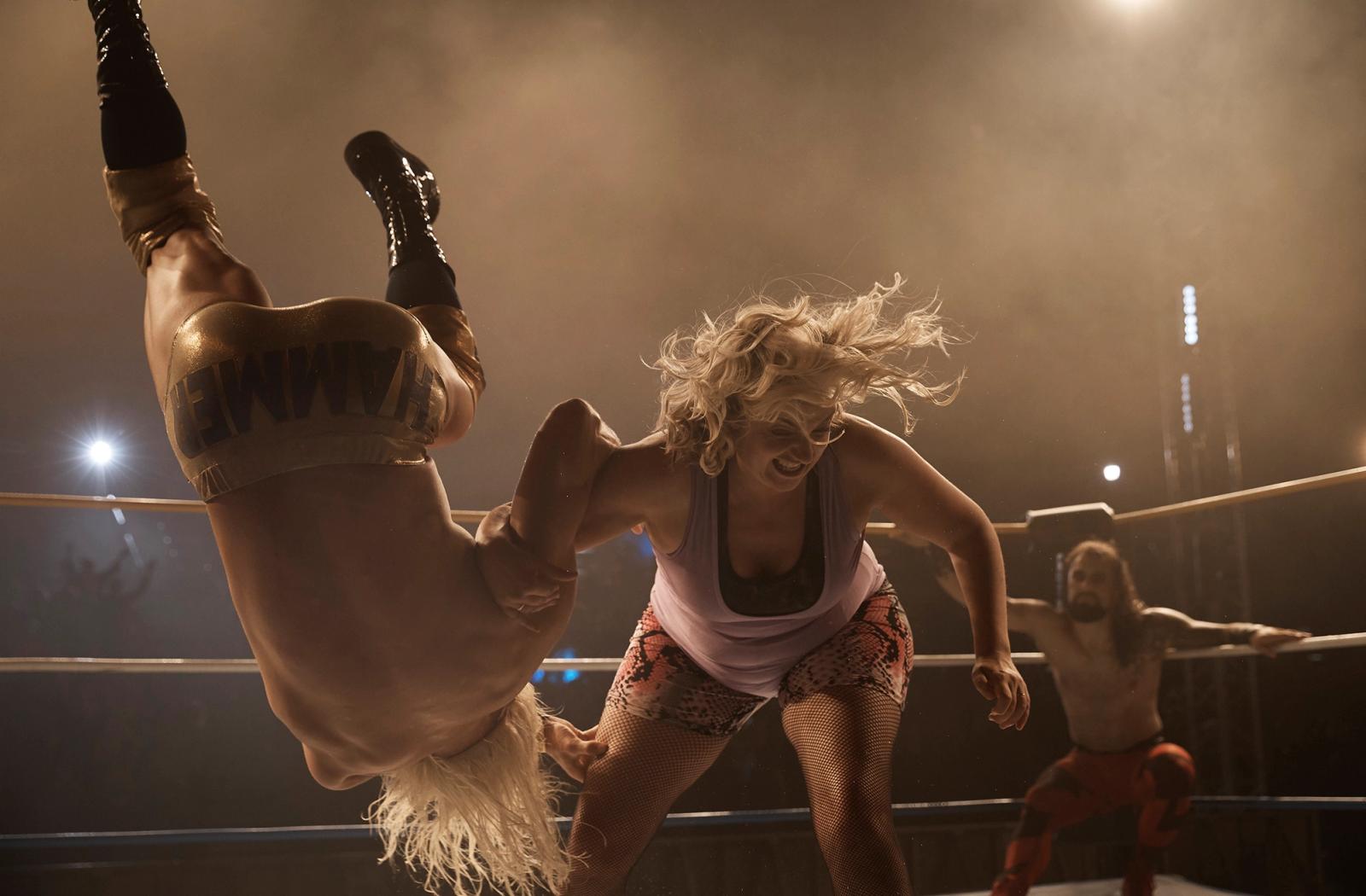 A female wrestler in a pink tank top and leopard-print shorts is being thrown by a male wrestler in a gold and black wrestling costume, with a crowd in the background.