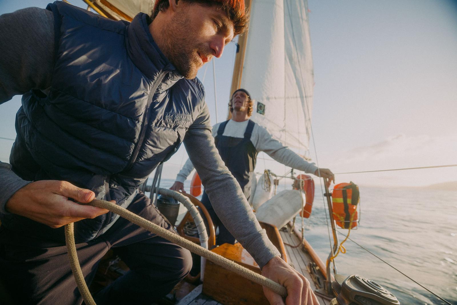 Two men on a sailboat, one holding a rope, with a white sail and a life ring on the deck, in a calm sea with a clear blue sky.