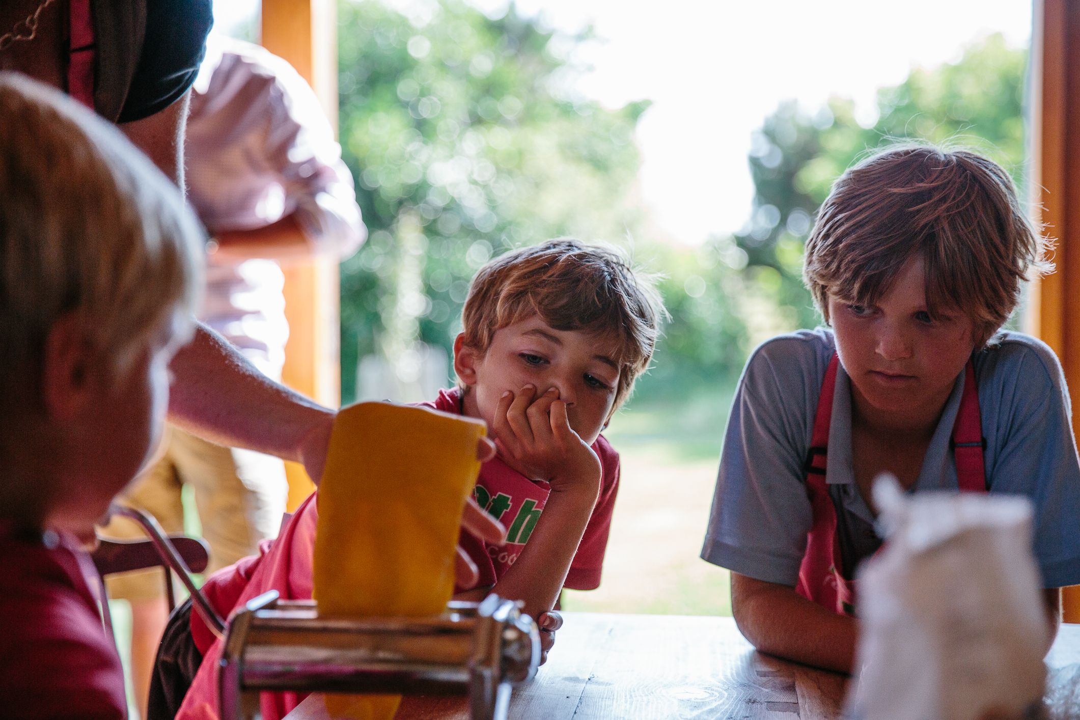 A group of children are learning to make pasta with a chef in a kitchen setting, with a pasta machine in the foreground.