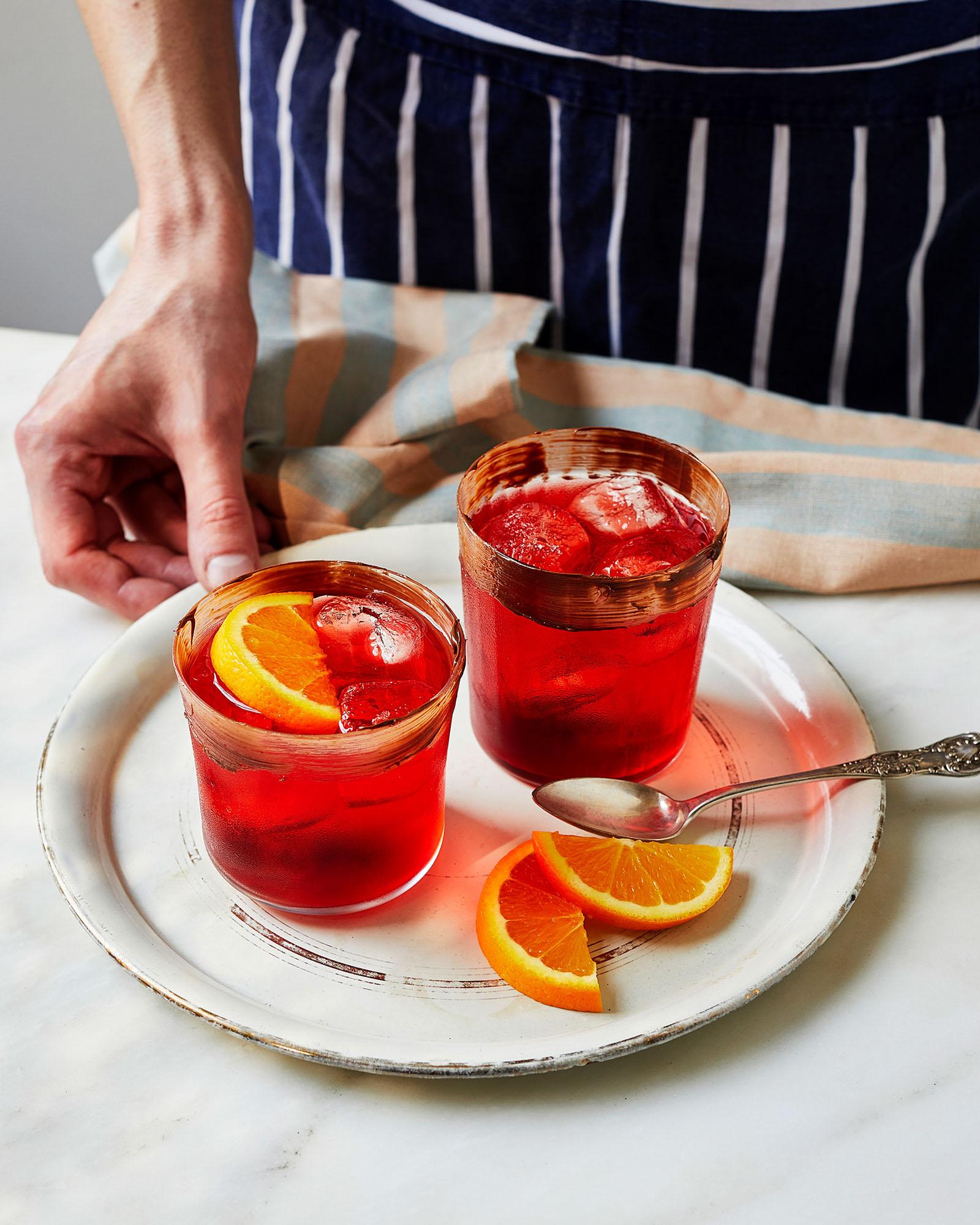A person in a blue and white striped apron holds a tray with two glasses of red liquid, garnished with orange slices and ice.