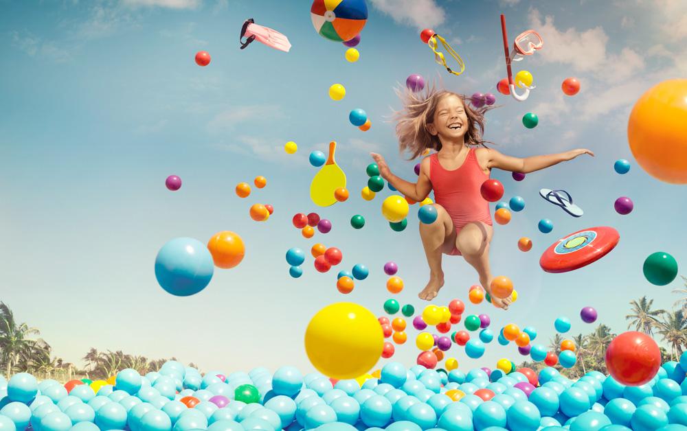 A young girl in a red swimsuit jumping into a ball pit filled with blue balls, surrounded by various beach toys and accessories.