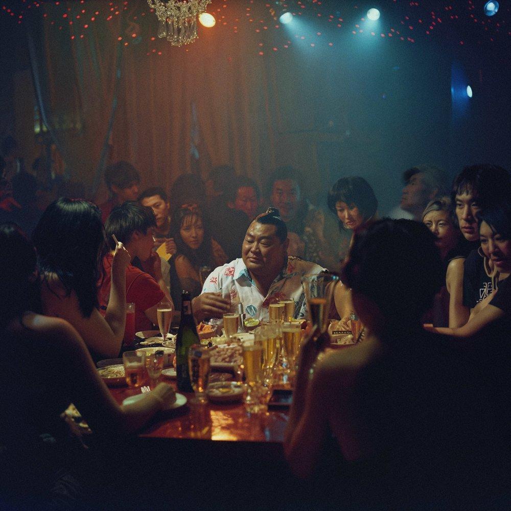 A crowded nightclub with patrons sitting at a bar, surrounded by neon lights and a DJ spinning music.
