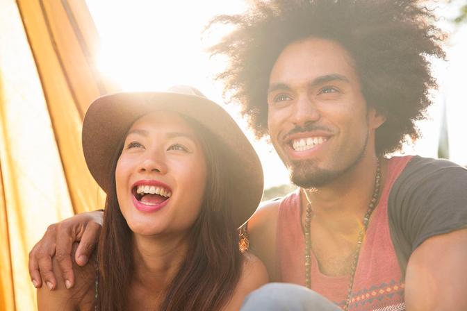 A young woman with a big smile and a wide-brimmed hat is laughing and leaning on a man with a beard and a tank top, both with their arms around each other.