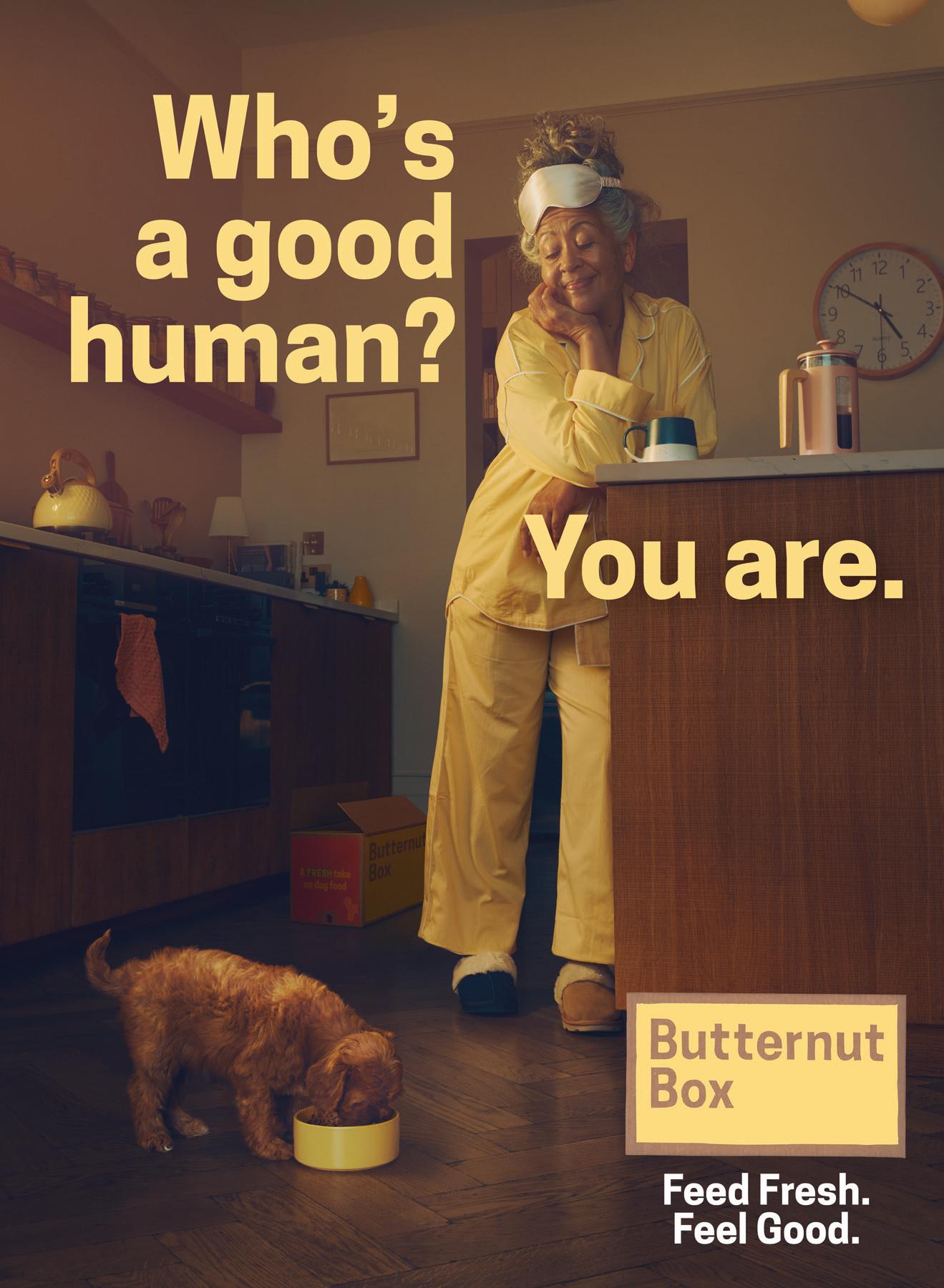 A woman sits at a kitchen counter while a dog eats from a bowl on the floor.