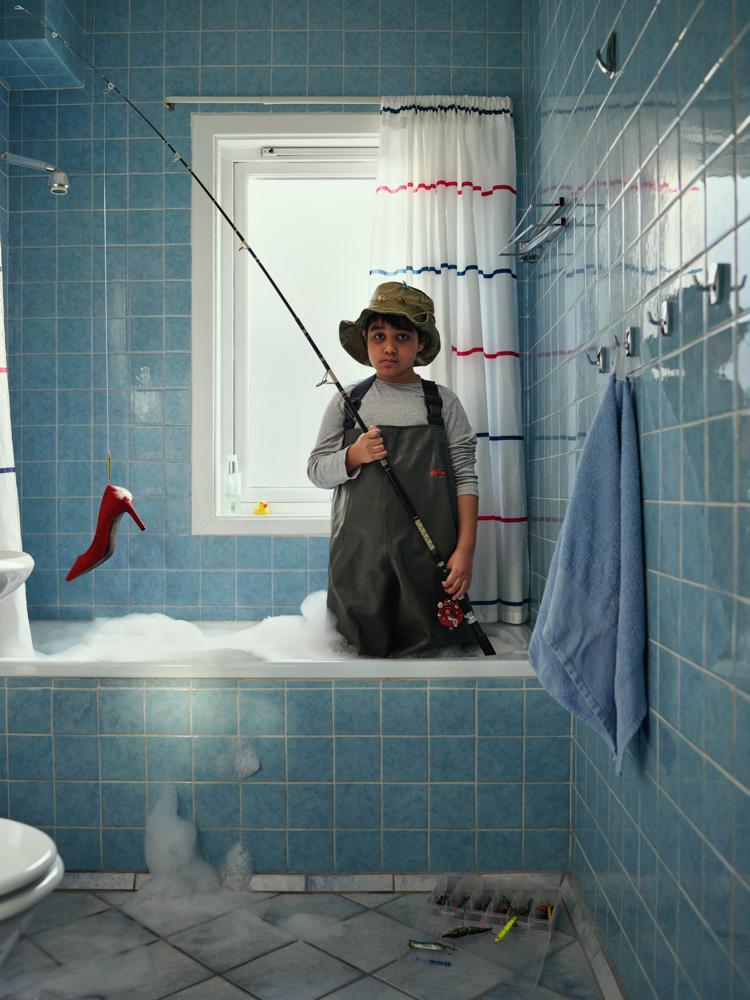 A young boy in a fishing wetsuit, holding a fishing rod, standing in a bathtub filled with soap bubbles, with a red high-heeled shoe hanging from the bathtub's faucet.