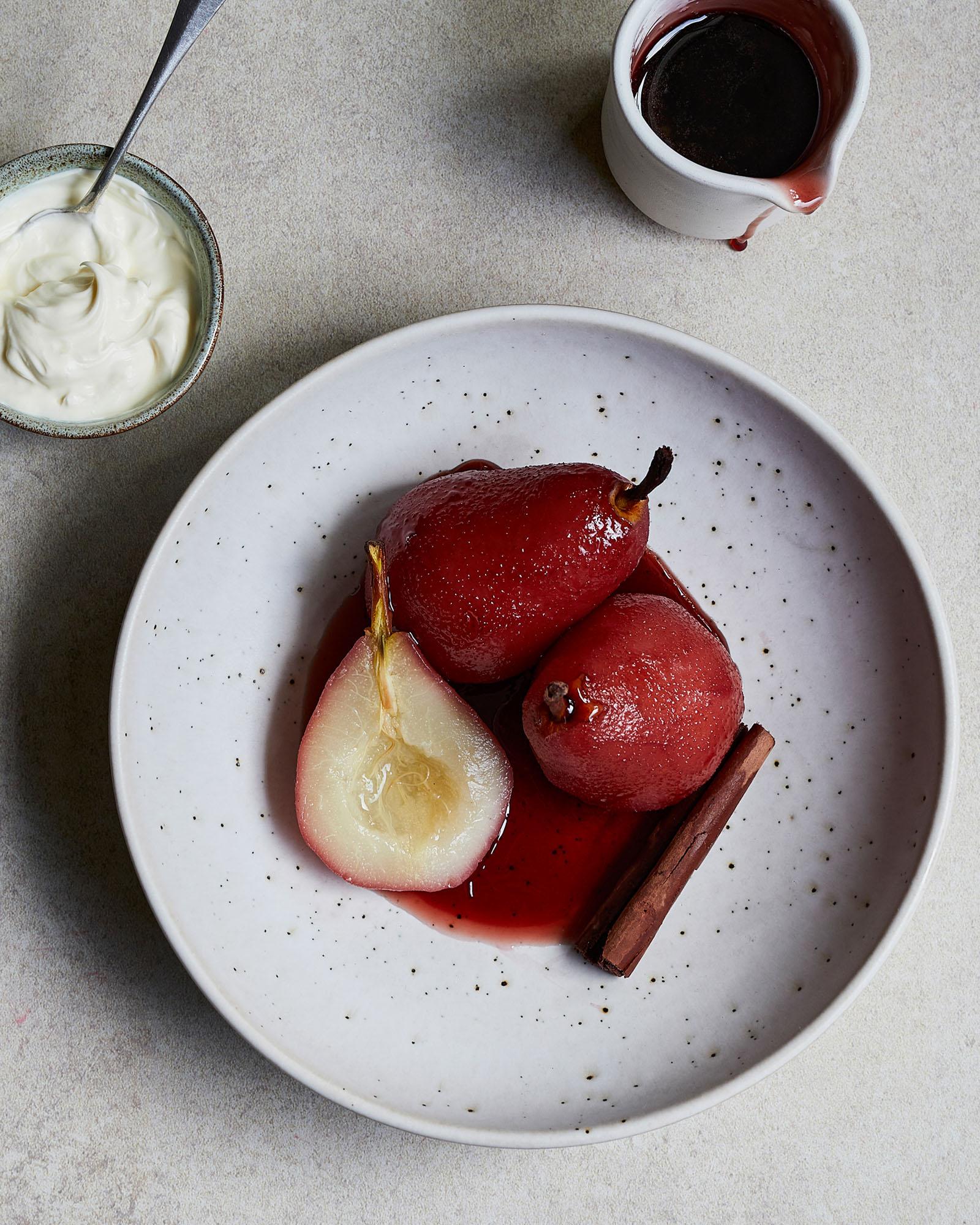 A plate featuring poached pears in red wine sauce with a cinnamon stick and additional pear slice garnish.