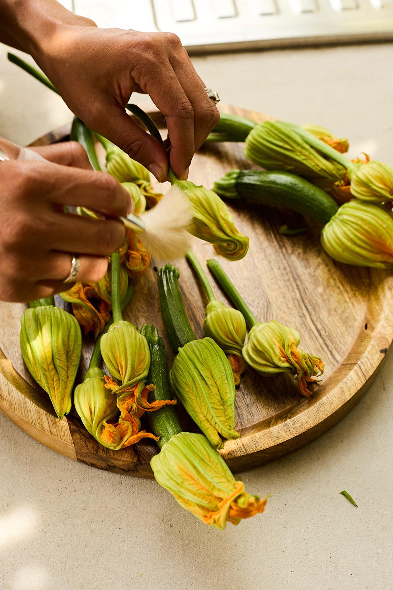 A person's hands are gently cleaning and preparing a variety of fresh, green, and vibrant flowers, including zucchini flowers and other types of flowers, on a wooden table.