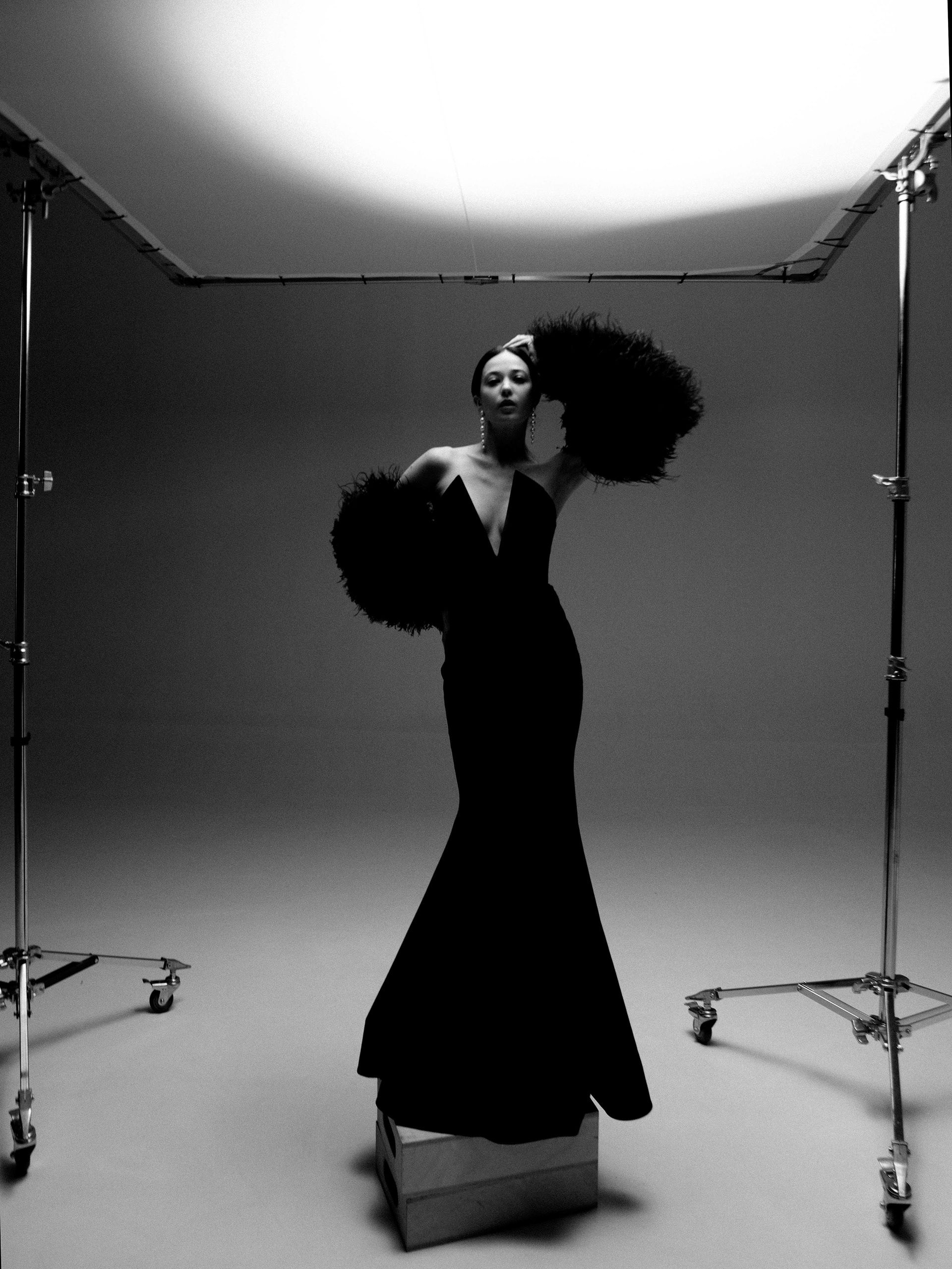A woman in a long, dark, strapless evening gown with a dramatic, flared train, posing in a studio with a large, white, circular light source above her head.
