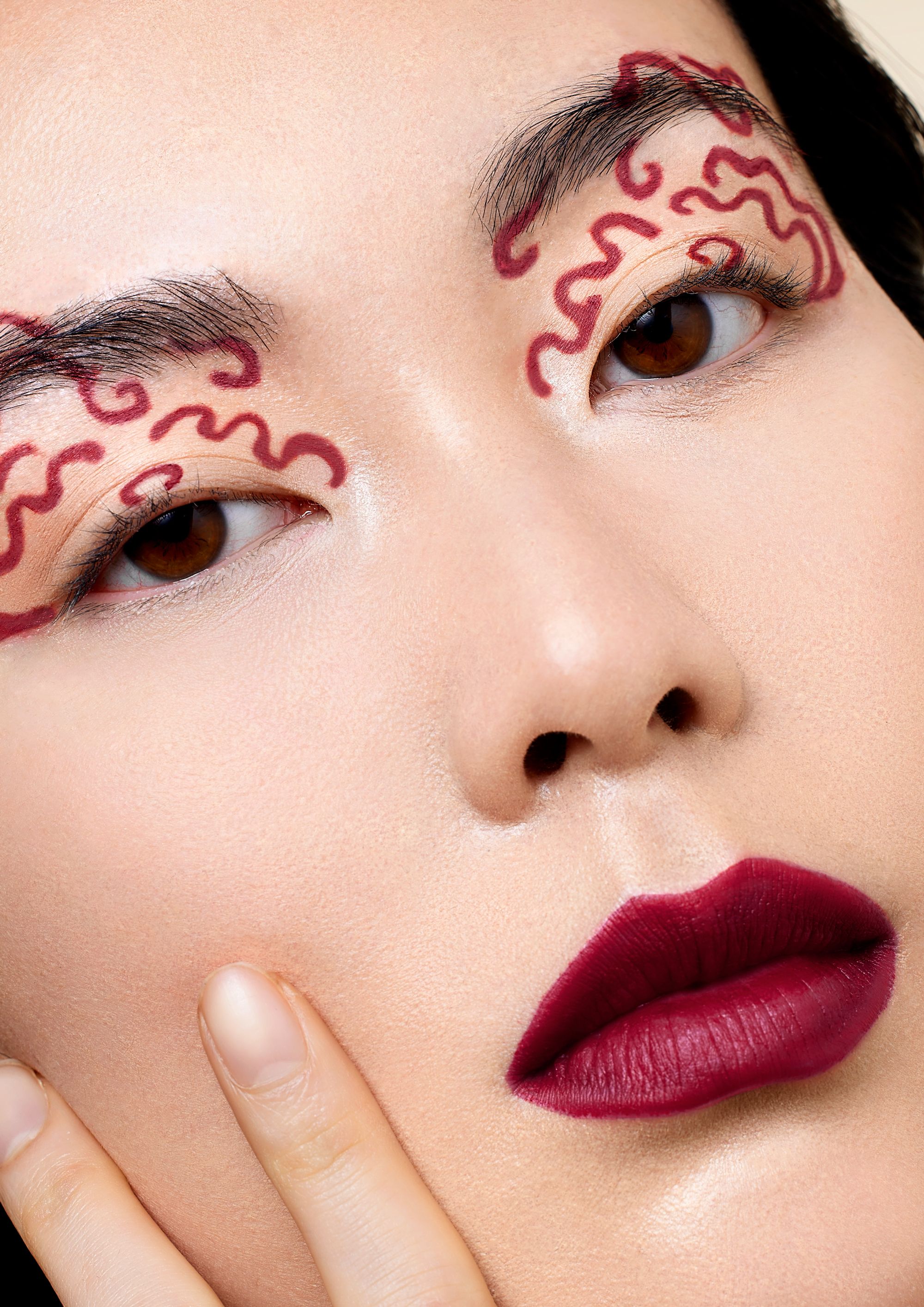 A close-up of a woman's face with intricate, red, hand-drawn designs on her skin, including her eyes, eyebrows, and forehead.