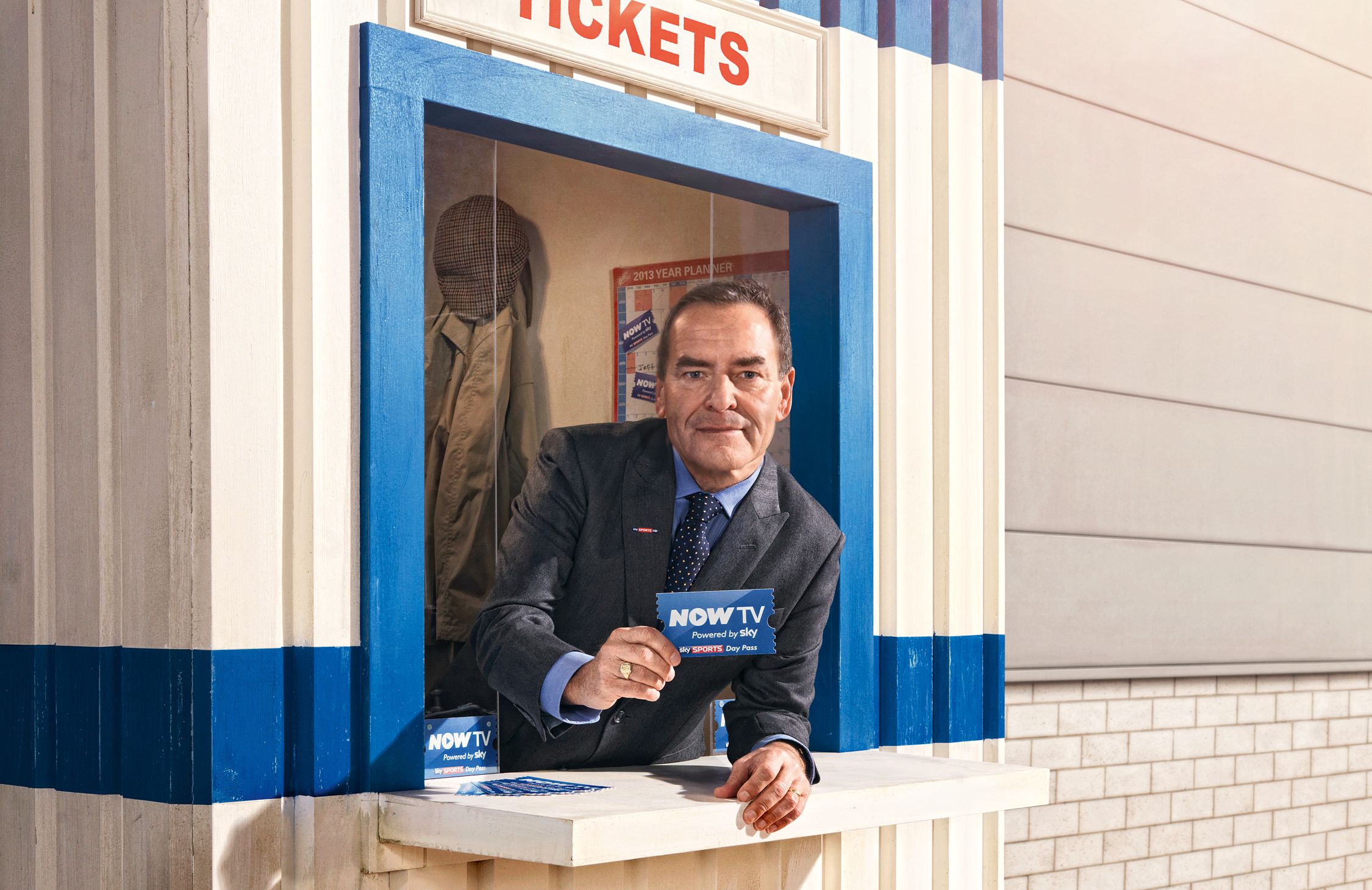 A man in a suit and tie standing behind a ticket booth, holding a ticket with the words 'NOW TV' on it.