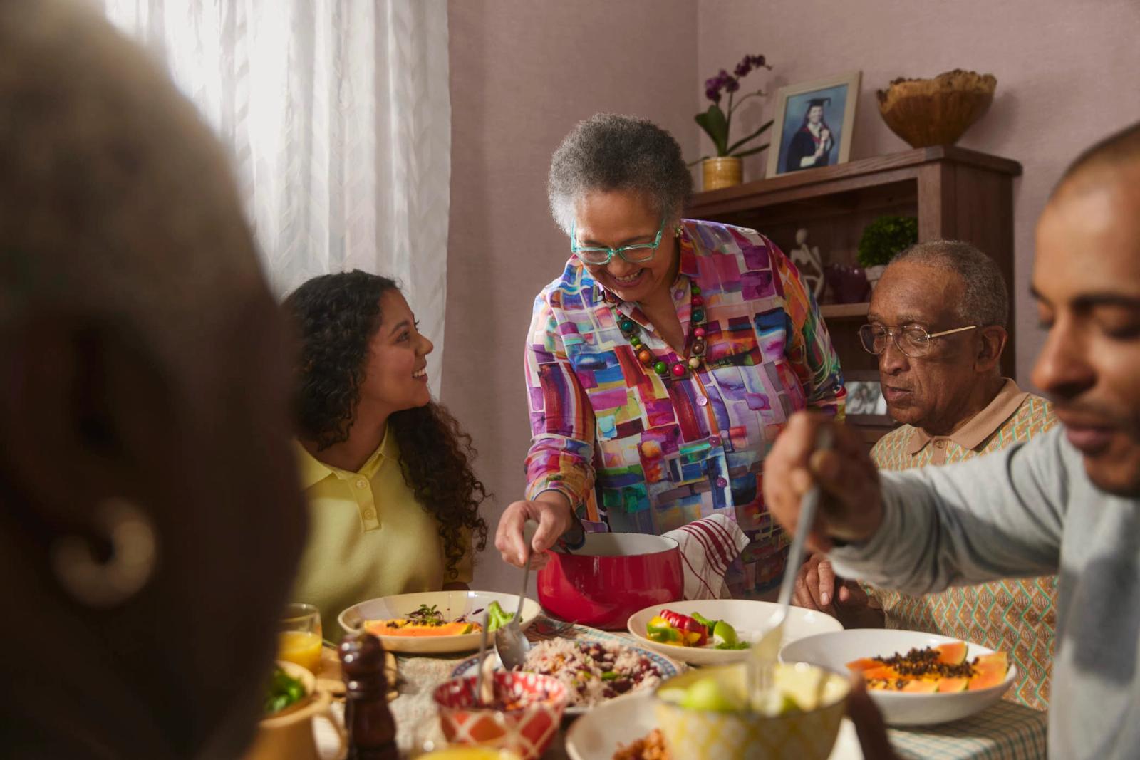 A woman with short gray hair and a colorful shirt is serving food to a group of people around a table. She is wearing a multicolored shirt with a geometric pattern and has a big.