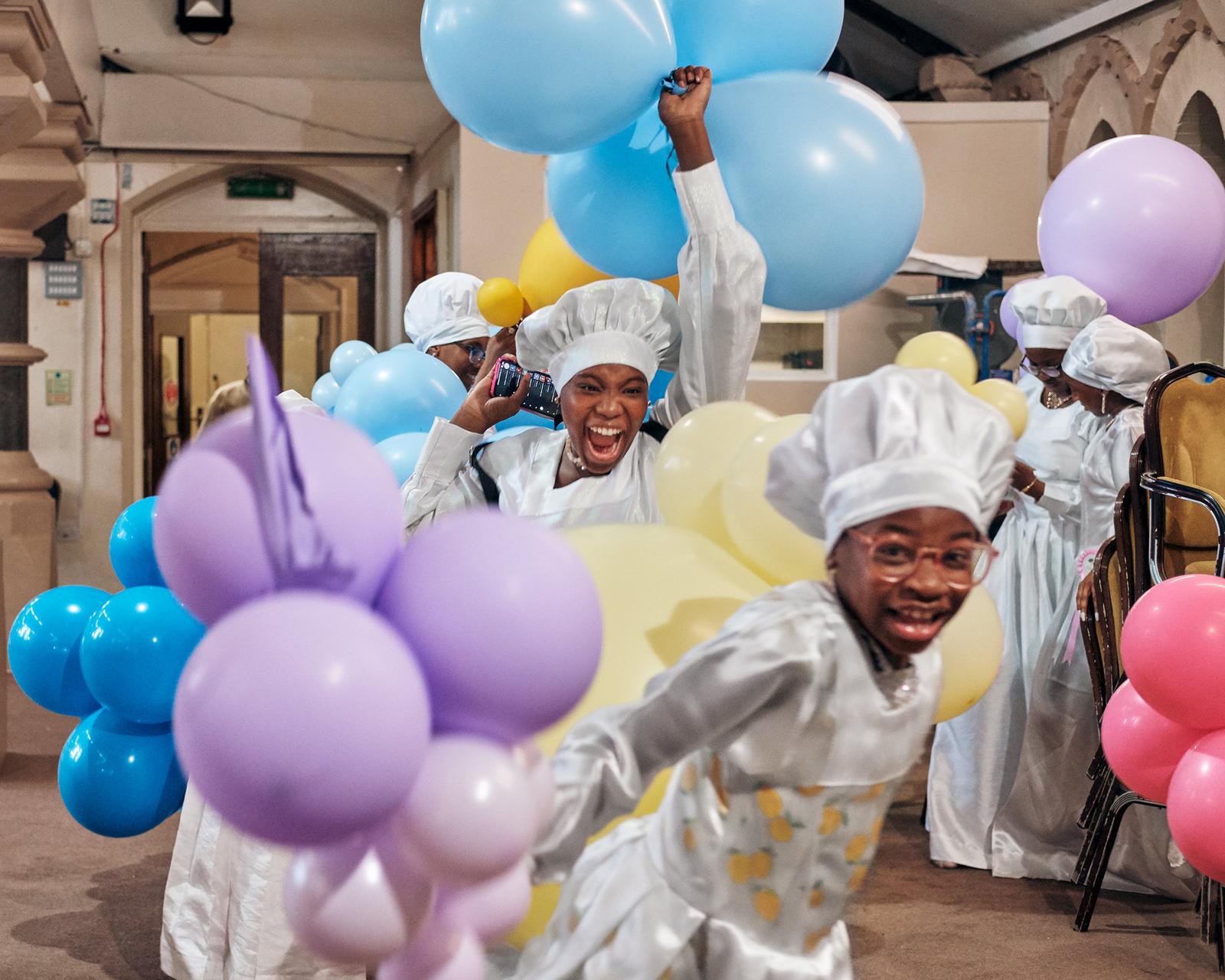 A group of people in white chef's uniforms and hats, surrounded by balloons and holding cameras, in a large, empty, industrial-style room with high ceilings and exposed pipes.