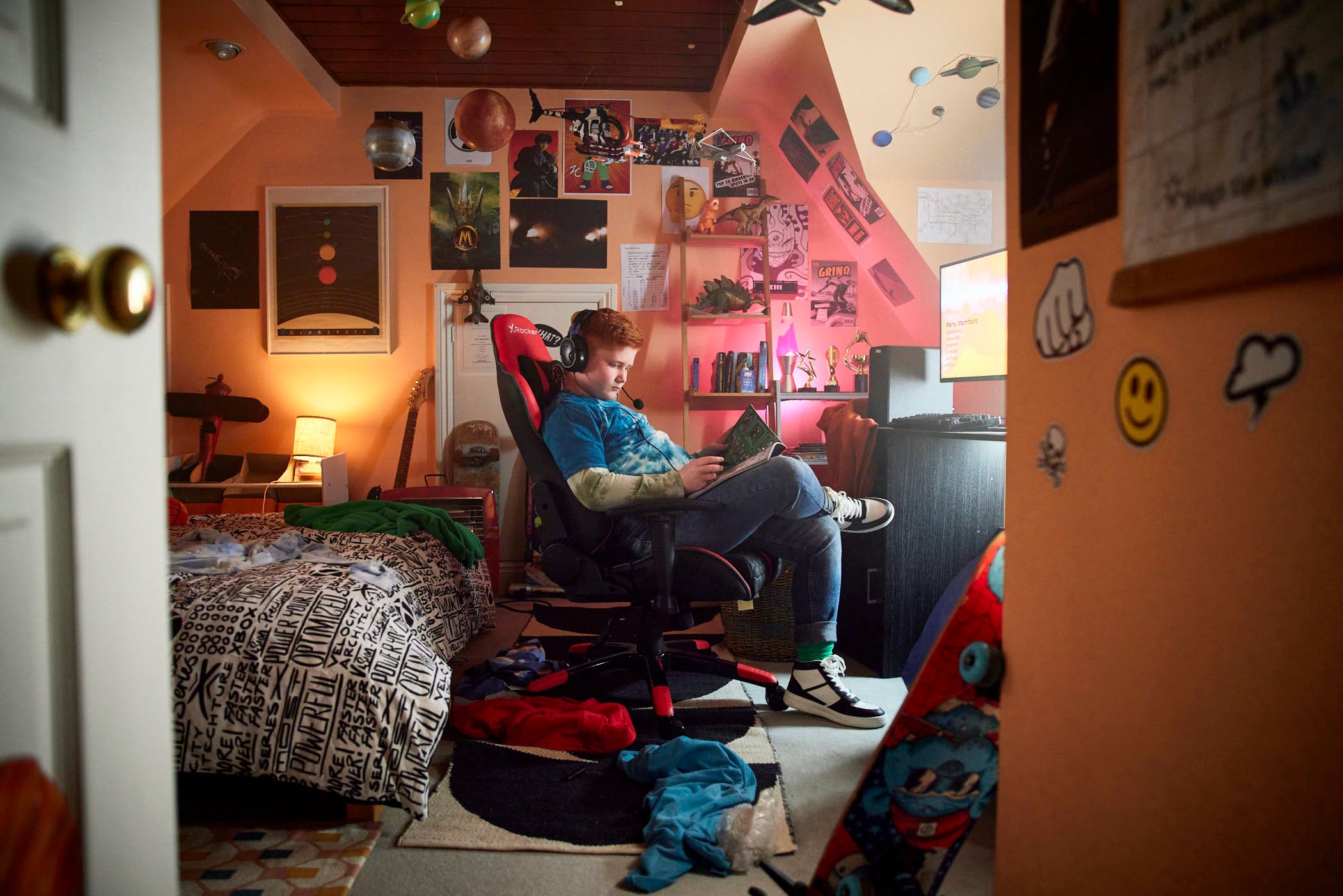 A young boy sits in a red gaming chair in a bedroom, engrossed in reading a book. He is wearing a blue tie-dye shirt, jeans, and white sneakers, and has headphones on, suggesting.