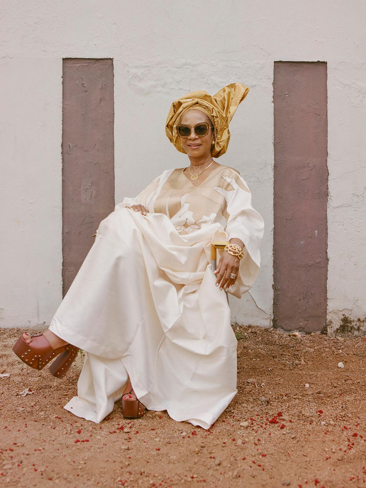 A woman wearing a white and gold traditional Yoruba attire, sitting on the ground with her legs crossed, surrounded by a white wall with three brown vertical panels.