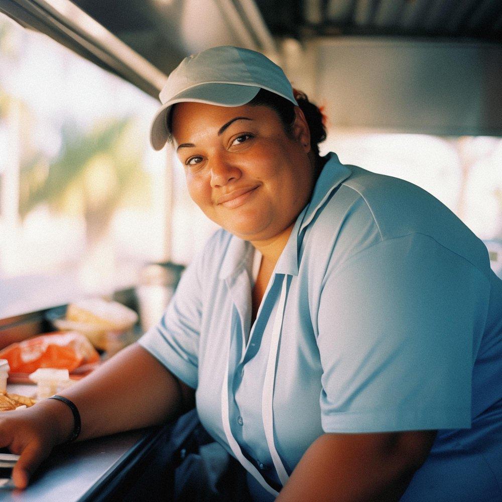 A woman in a white chef's hat and a blue apron is smiling and looking directly at the camera, standing in a kitchen with a counter and a sink in the background.