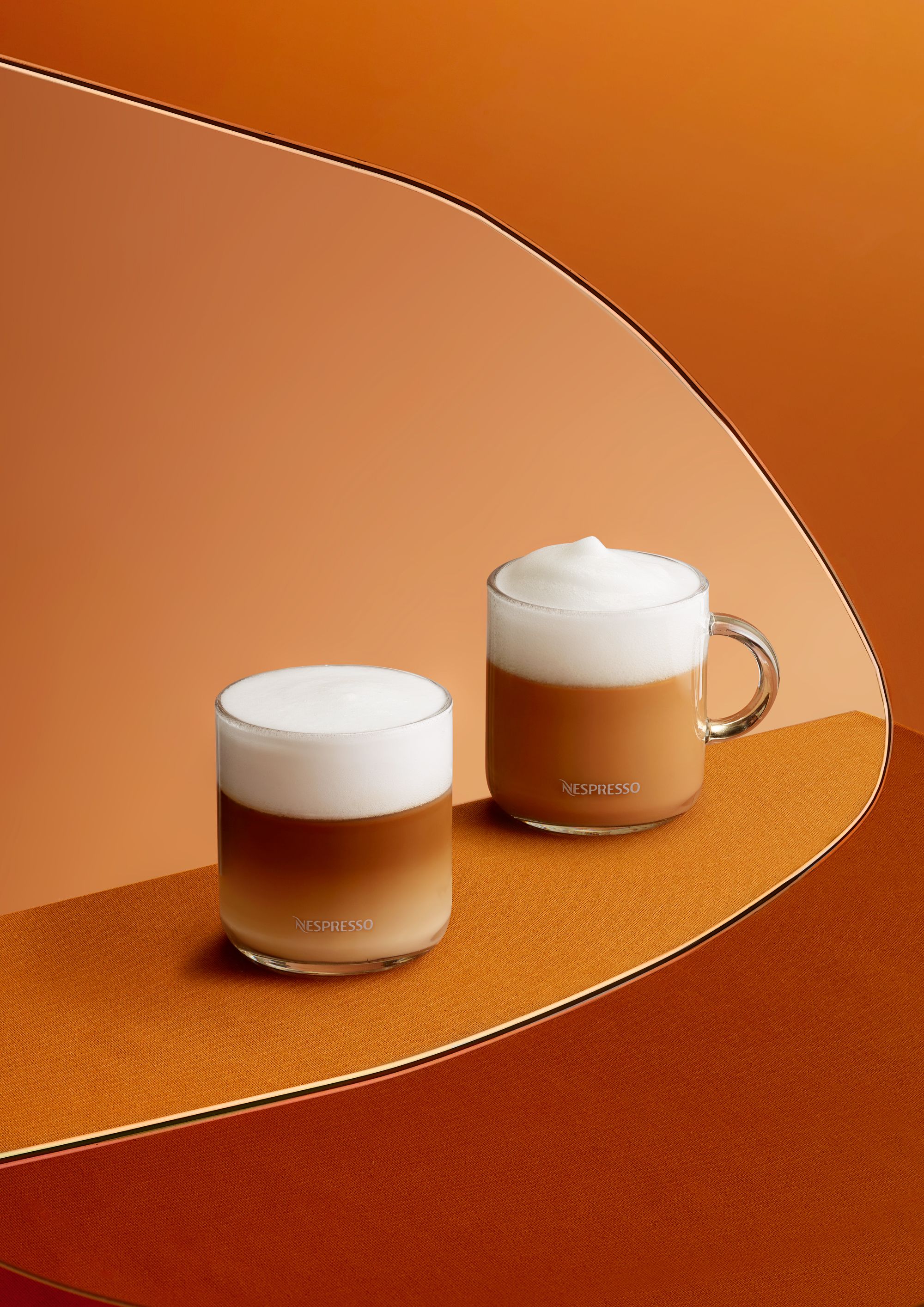 Two glasses of layered coffee drinks with a thick layer of foam on top, sitting on a curved, orange surface.