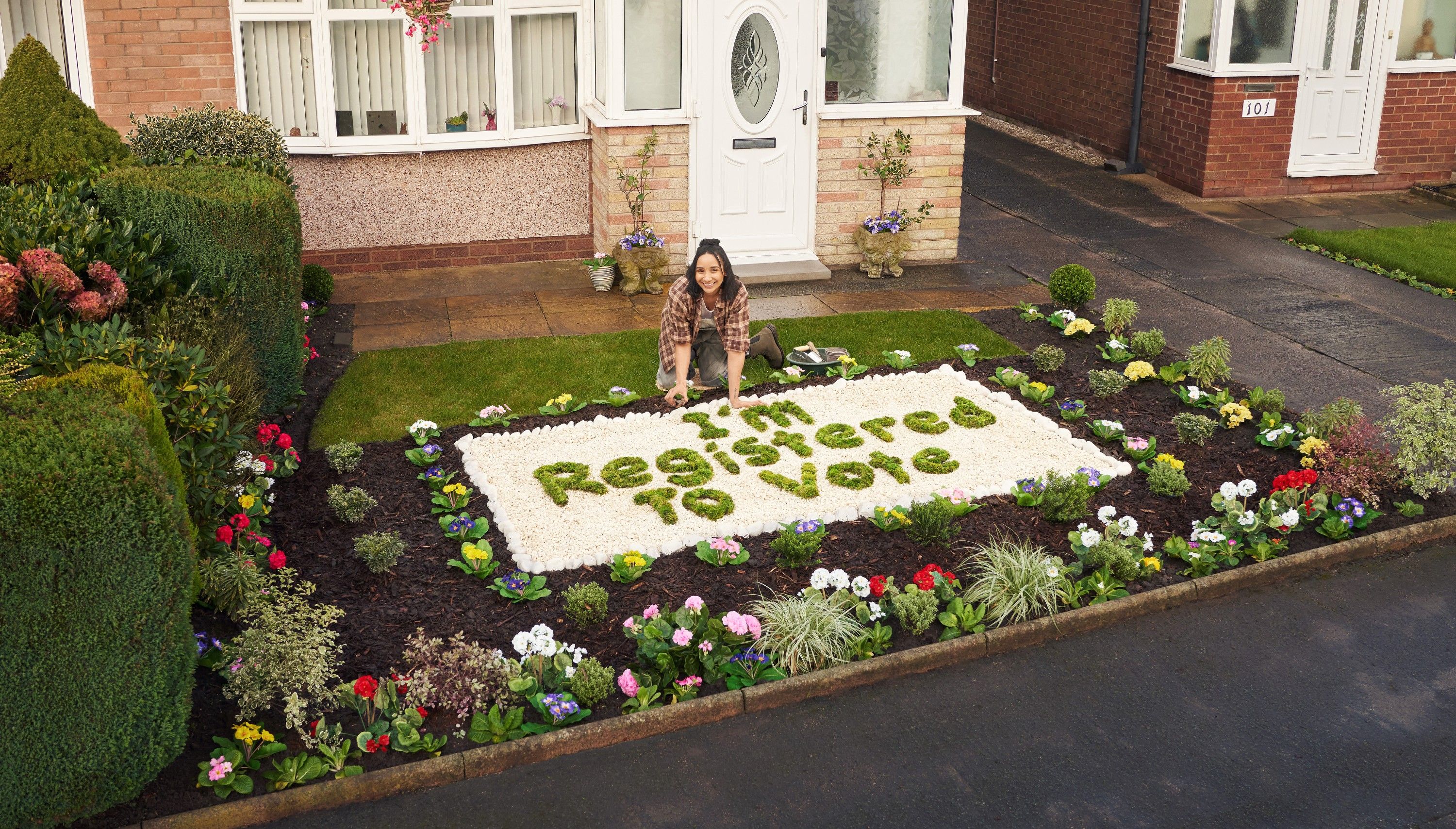 A woman kneels in a garden, surrounded by flowers and a large green sign reading 'I'm Registered to Vote'.