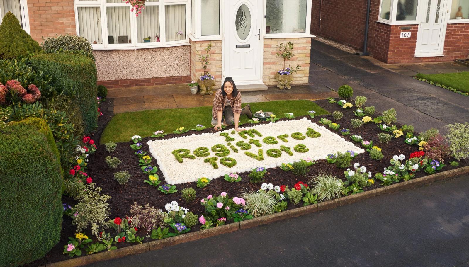 A woman kneels in a garden, surrounded by flowers and a large green sign reading 'I'm Registered to Vote'.