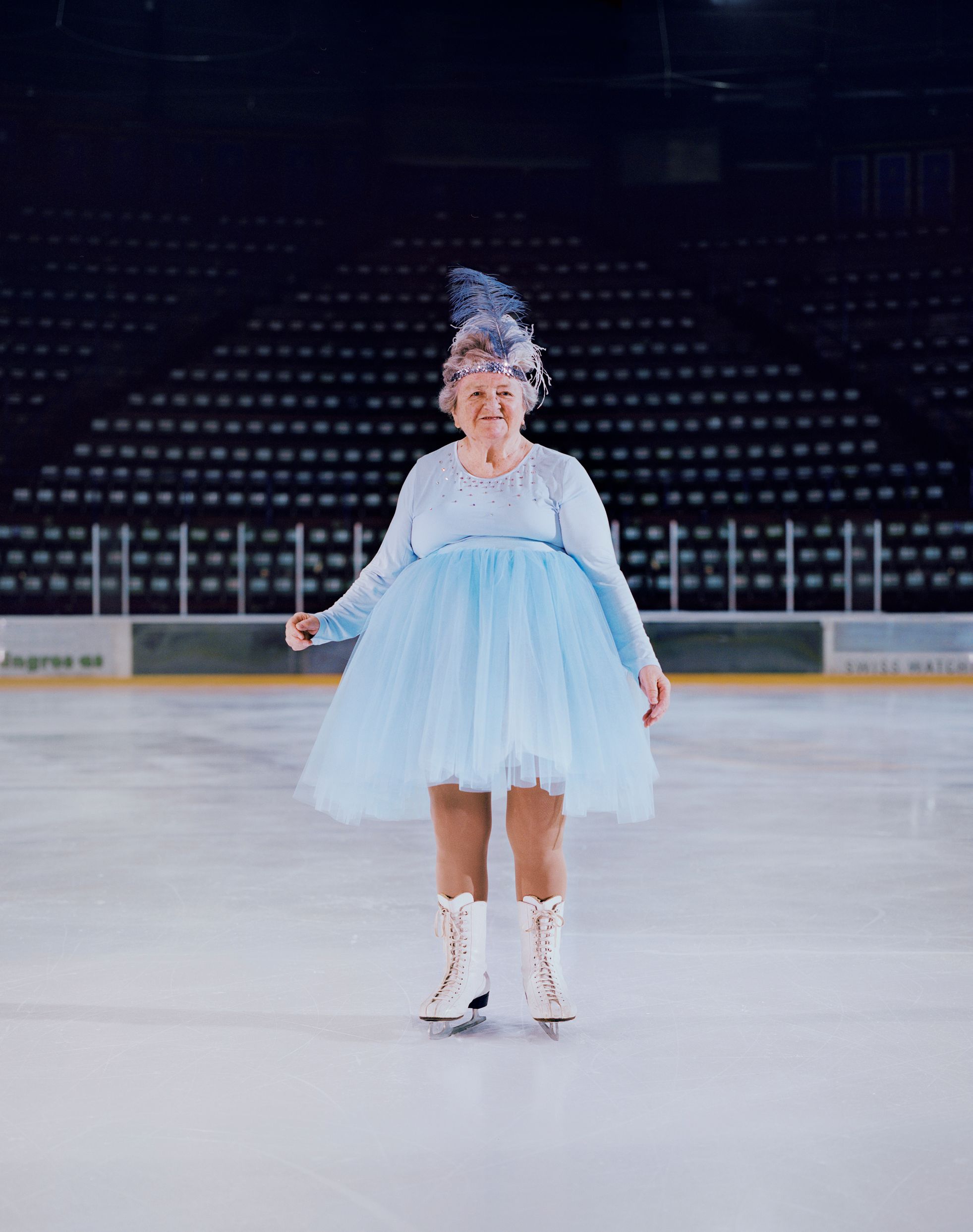 An elderly woman in a blue tutu and ice skates is standing on a frozen ice rink.