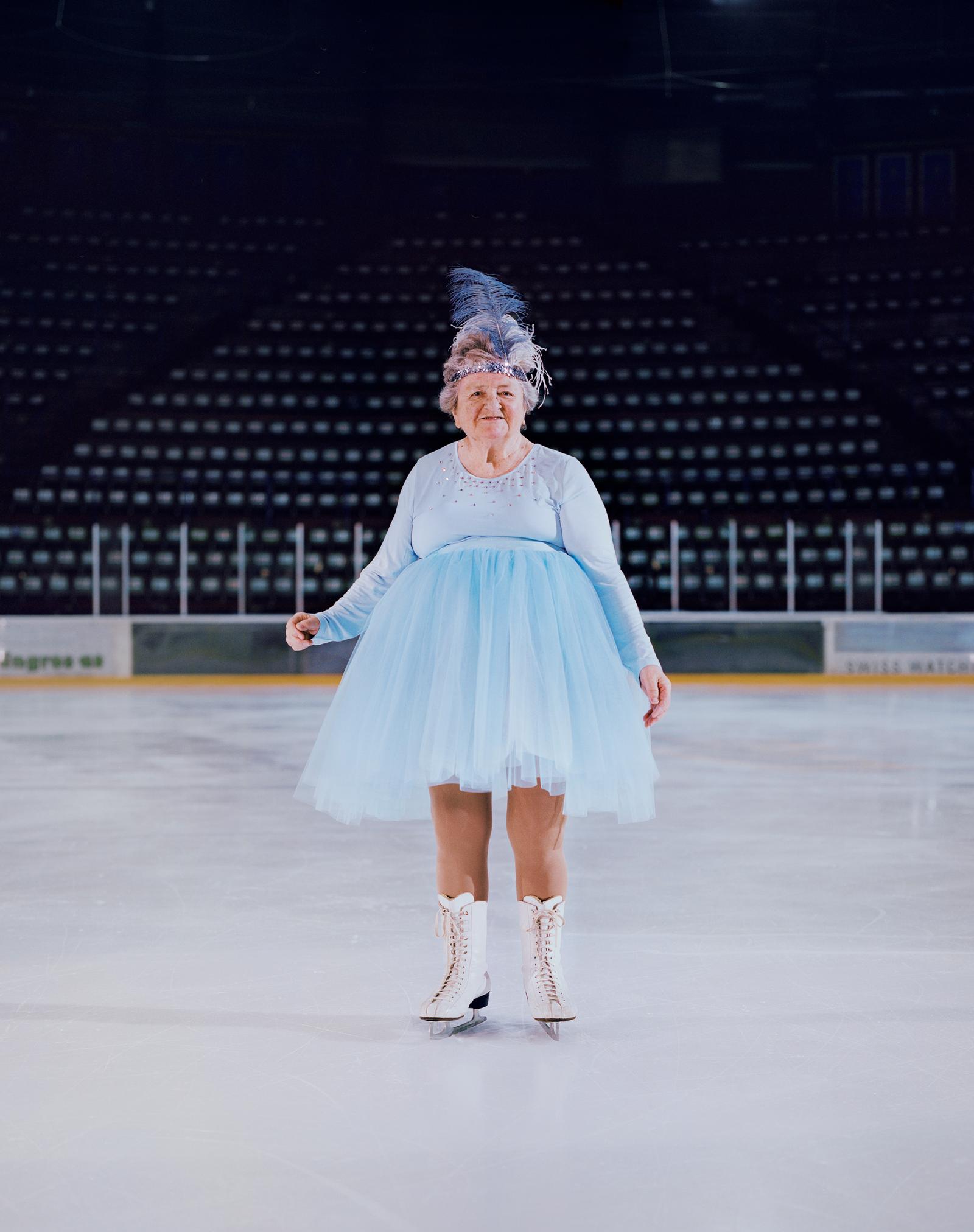 An elderly woman in a blue tutu and ice skates is standing on a frozen ice rink.