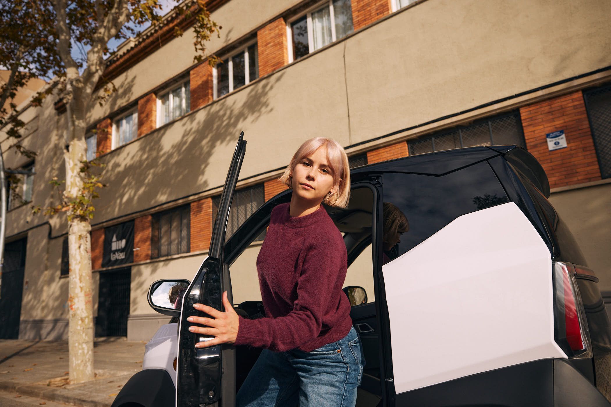 A woman with short blonde hair and a maroon sweater stands in front of a white and black car, her hand on the door handle. She looks directly at the camera with a serious.