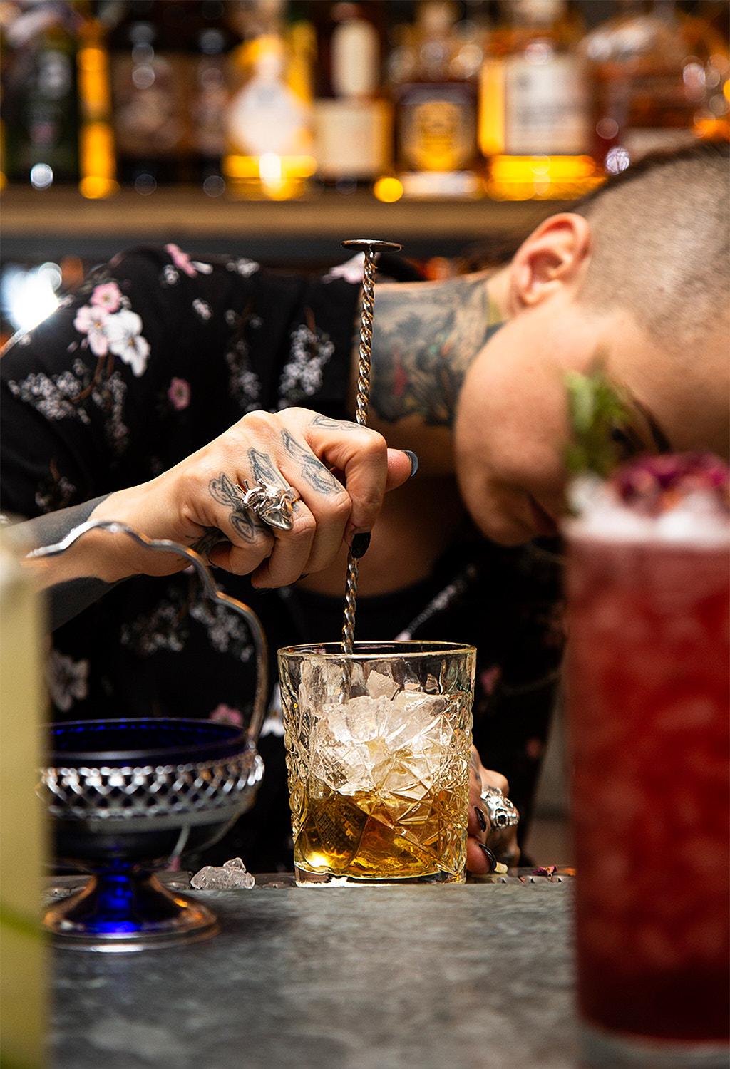 A bartender is expertly mixing a cocktail with a metal cocktail shaker, surrounded by various bottles and ingredients on a counter in a dimly lit, upscale bar.