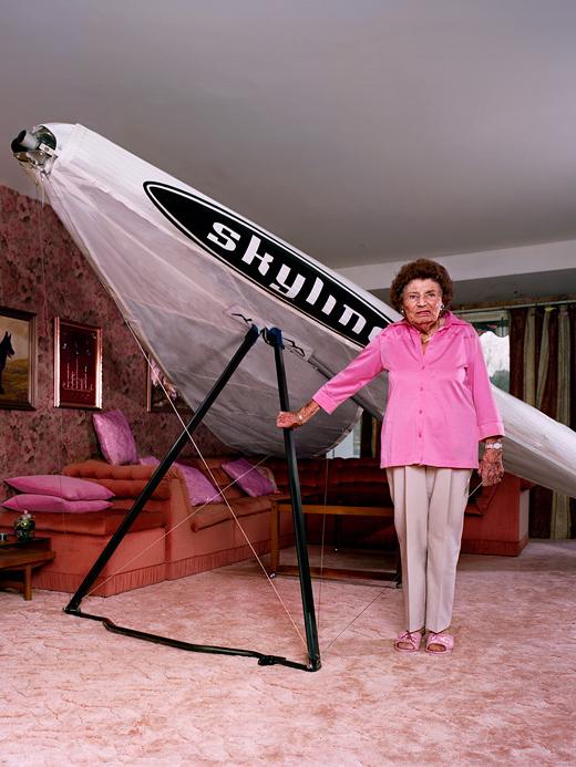 A woman in a pink shirt and beige pants stands in front of a large, white, inflatable SkyMall airship in a living room with pink and white furniture and decorations.
