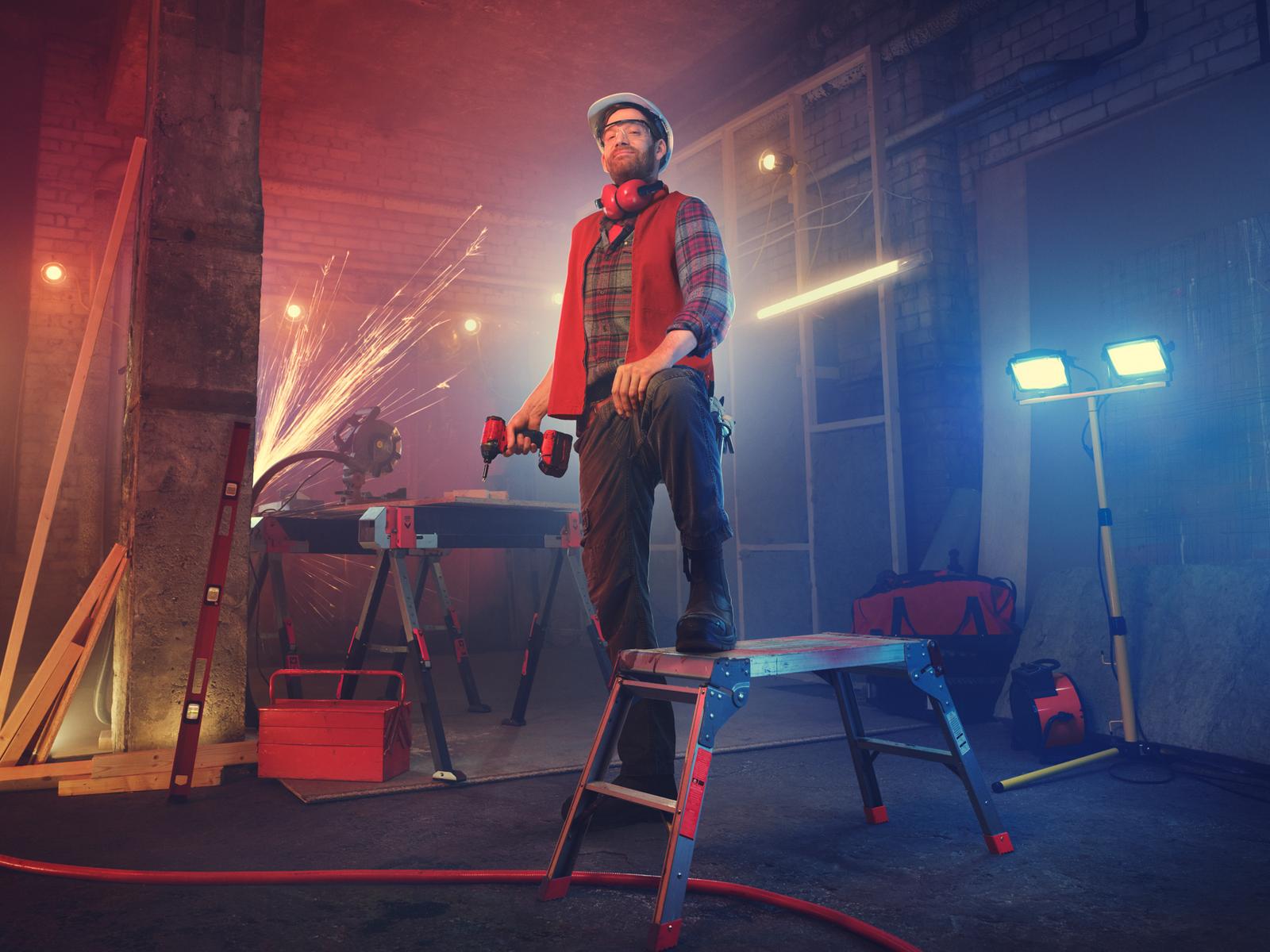 A man in a hard hat, plaid shirt, and jeans, holding a drill and standing in a workshop with various tools and equipment.
