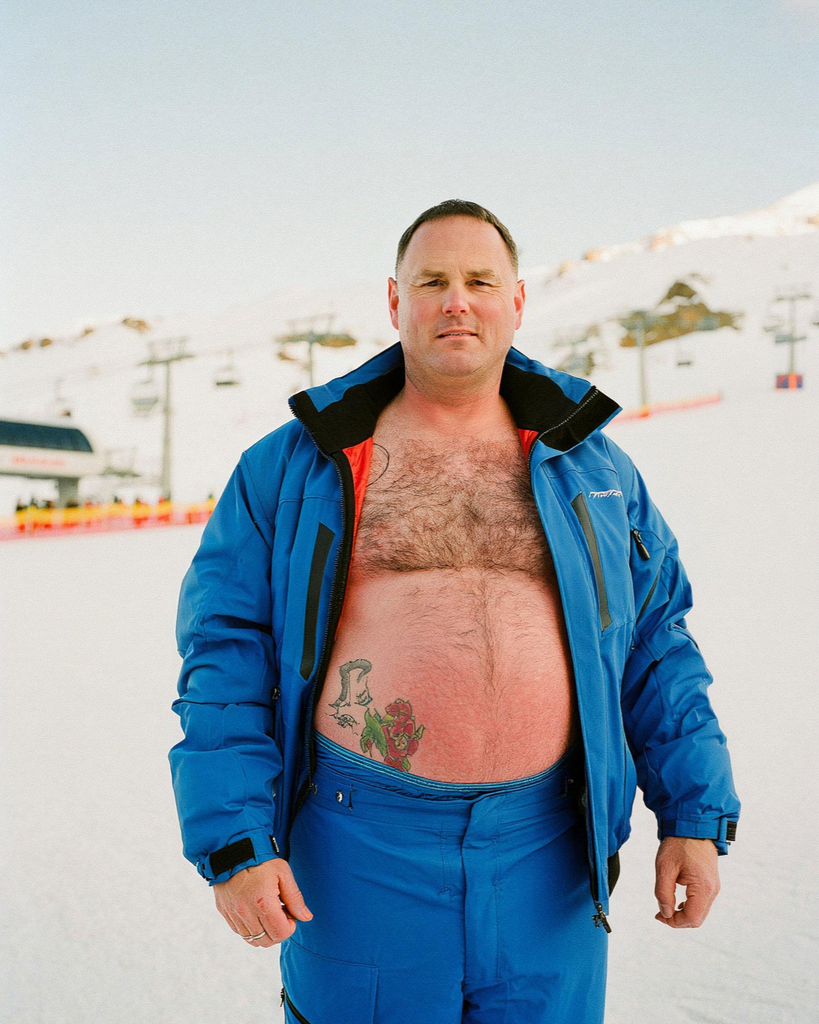 A man in a blue ski jacket and blue snow pants, with a large, colorful tattoo of a rose on his lower abdomen, standing in a snowy, mountainous landscape.
