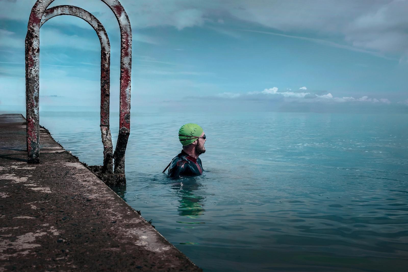 A man in a green swim cap and goggles stands in the water, gazing out at the horizon. He is wearing a wetsuit and has a towel around his neck, and his arms are crossed over his.