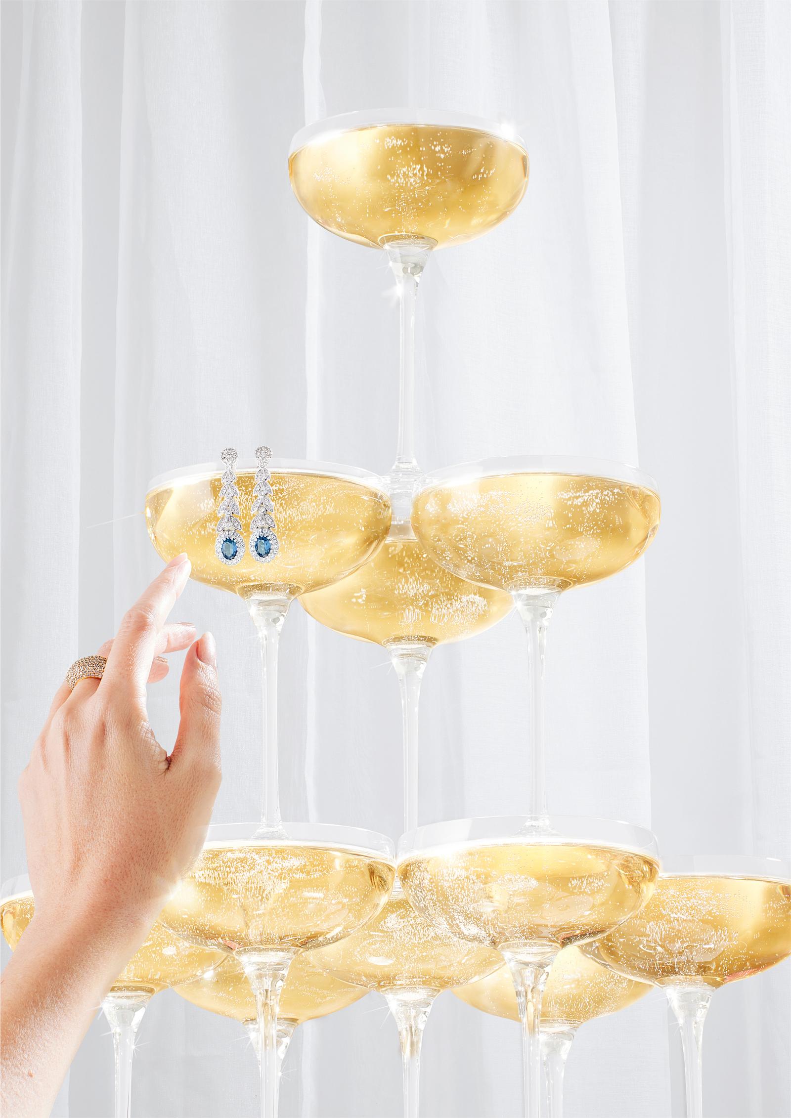 A hand reaching for a champagne glass with a diamond earring hanging from the rim, amidst a stack of champagne glasses.