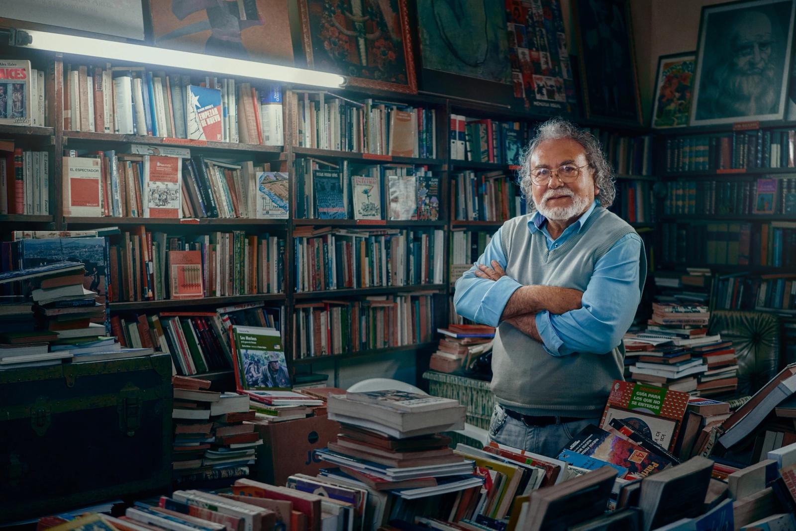 A man stands in a room filled with books and art, arms crossed.