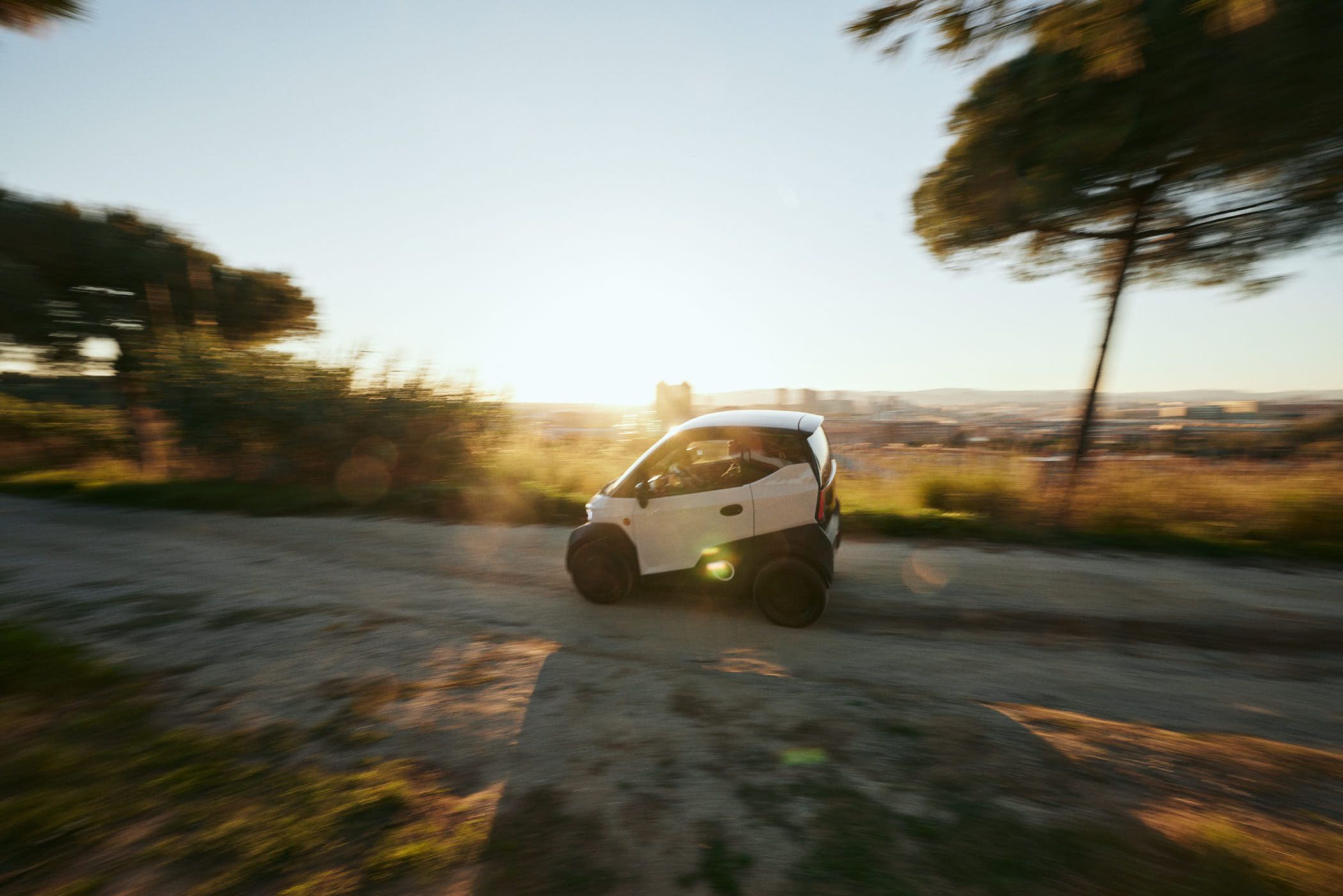 A white, compact car drives down a dirt road at sunset, its headlights casting a warm, golden light on the surrounding trees and grass. The driver, partially visible through the.