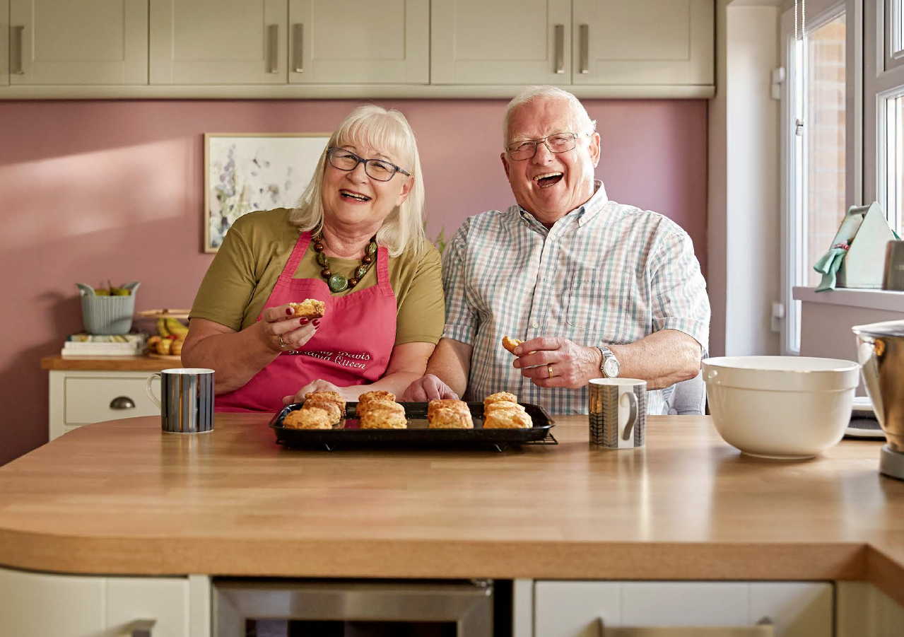An elderly woman and man, both with white hair, sitting at a kitchen counter, smiling and holding a tray of freshly baked scones.