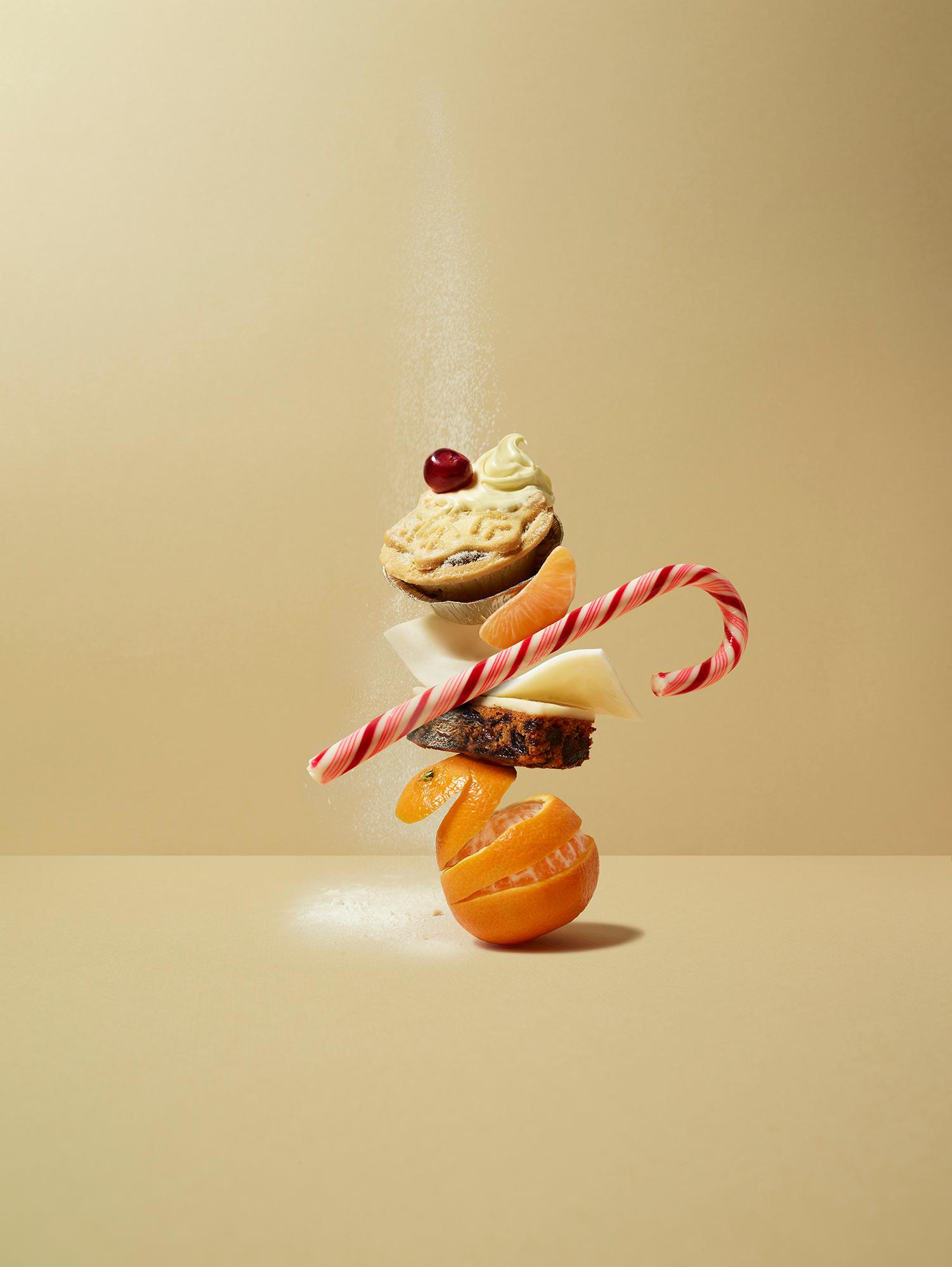 A stack of various foods, including a mince pie, candy cane, orange, and cheese, arranged in a precarious balance. The foods are suspended in mid-air, with a fine layer of flour.