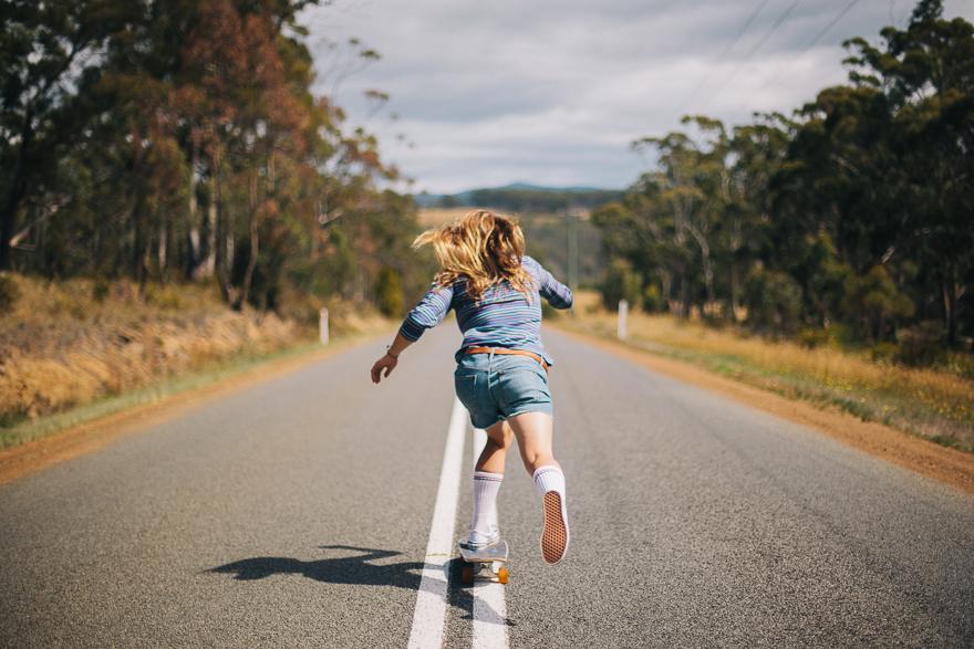 A young woman with long blonde hair is skateboarding down a long, straight, rural road, wearing a blue and white striped shirt, denim shorts, white socks, and sneakers.