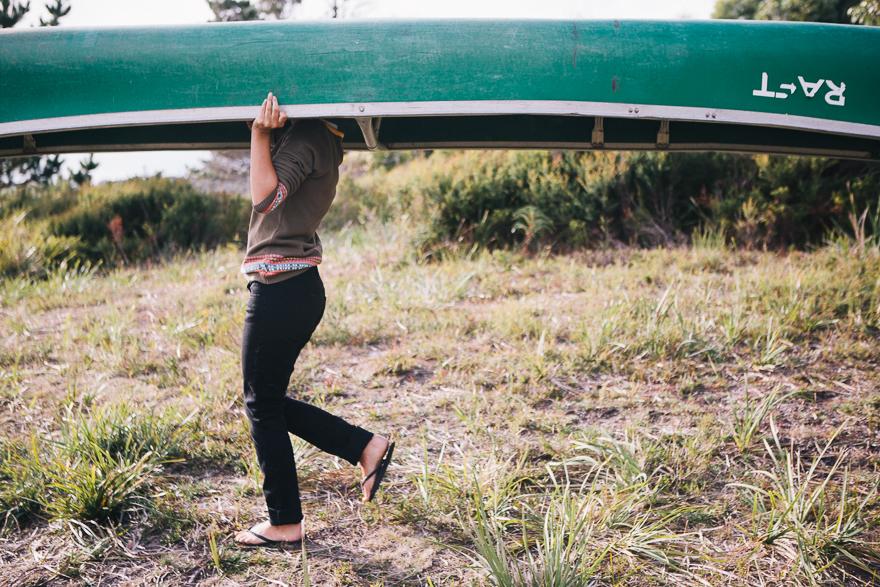 A woman carrying a green canoe on her head, walking through a grassy field.