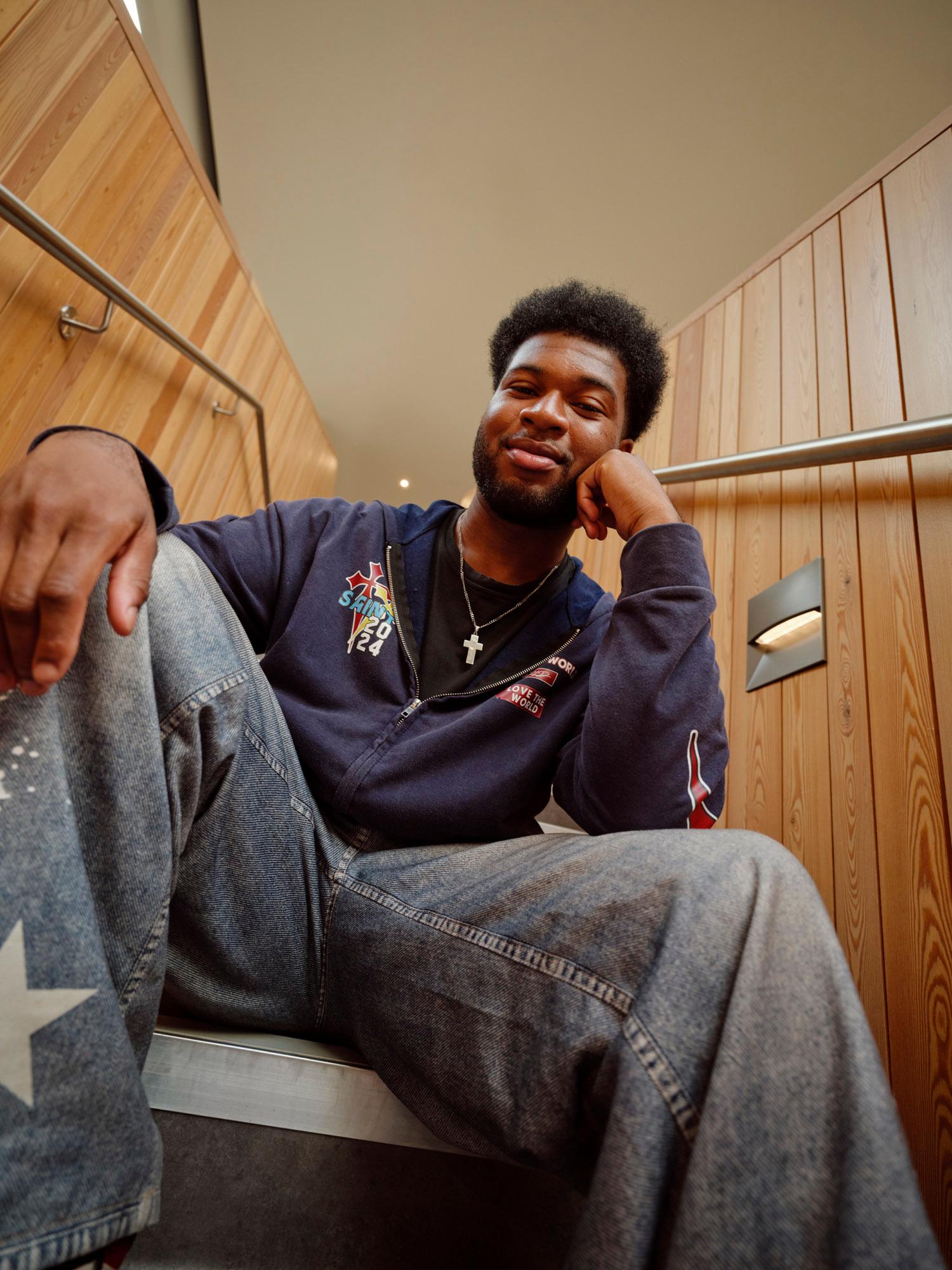 A man sits on a metal bench with his legs stretched out, wearing a blue hoodie with a red and yellow logo, jeans, and a silver necklace with a cross pendant.