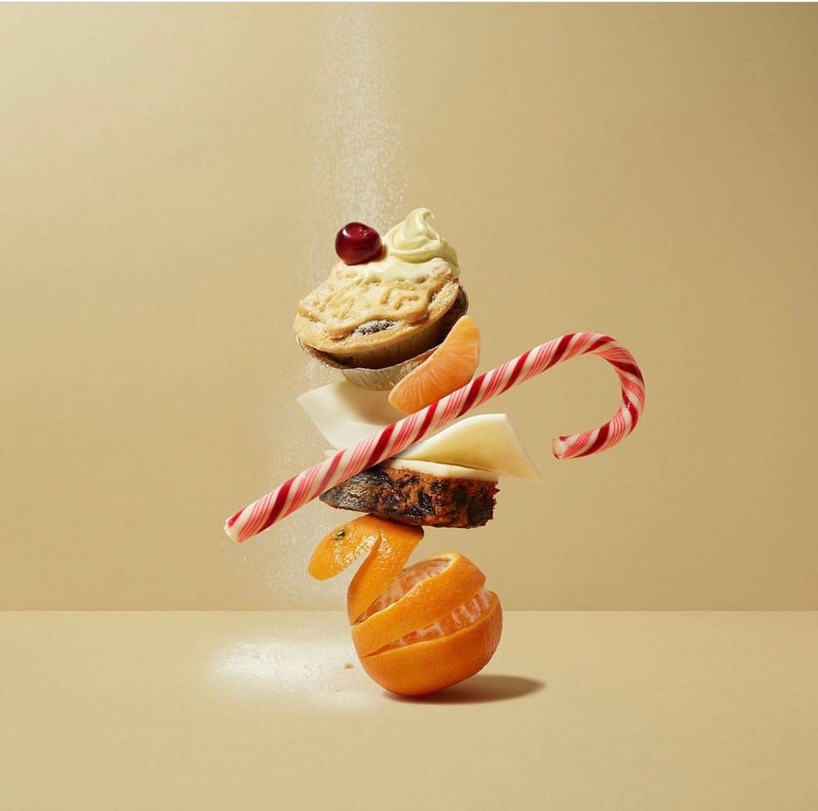 A stack of various Christmas treats, including a mince pie, a candy cane, an orange, and a piece of cheese, all arranged in a precarious and artistic way.