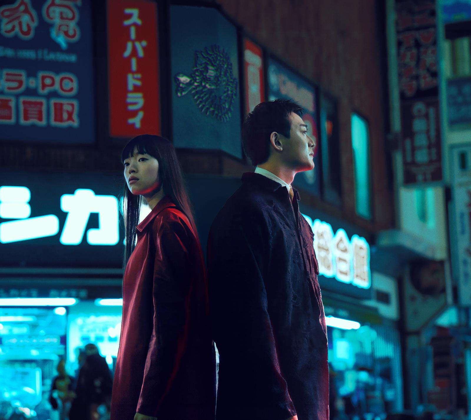 A man and a woman standing back-to-back, looking out over a city at night.