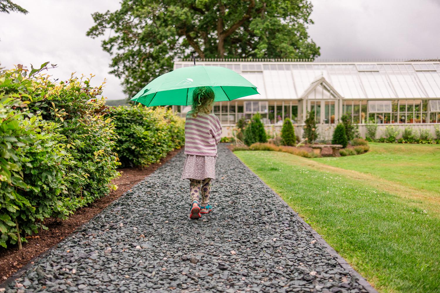 A young girl walking down a path in a garden, holding a green umbrella.