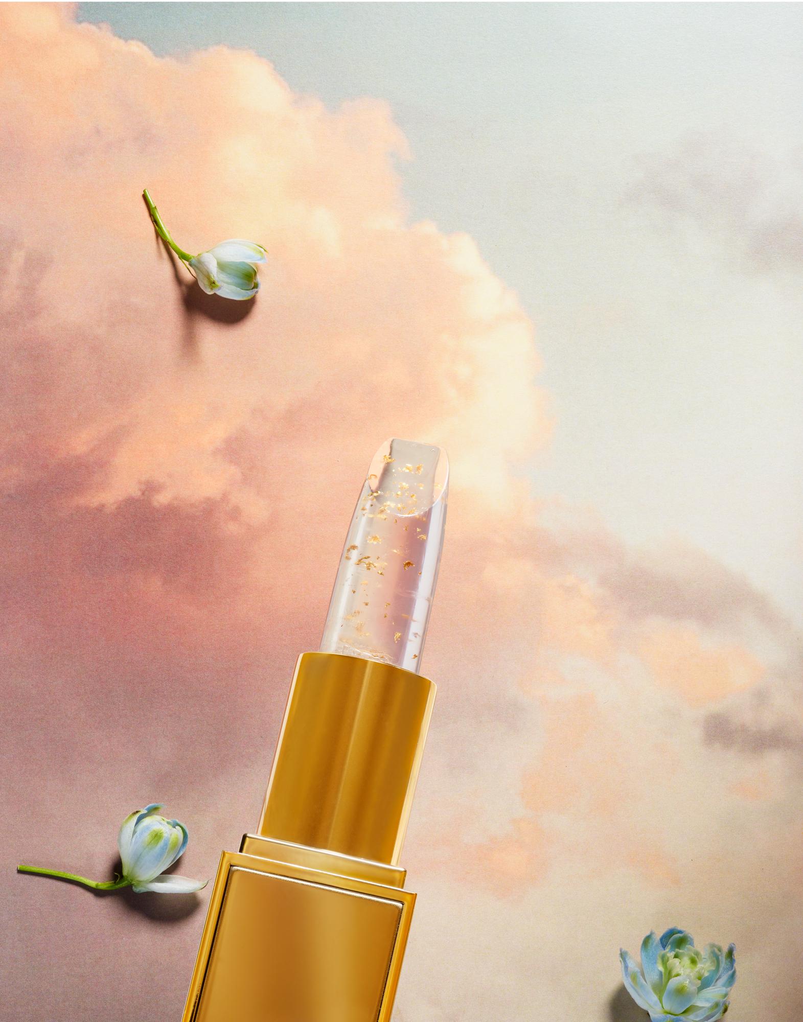 A gold lipstick with a clear, sparkly, gold-leaf-like substance inside, against a background of a pink and blue sky with clouds.