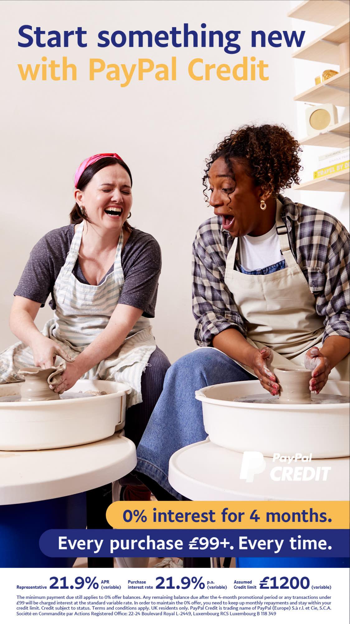 Two women in a pottery studio, one with dark hair and one with curly hair, both wearing aprons and laughing as they work on a pottery wheel. The woman with dark hair is creating.