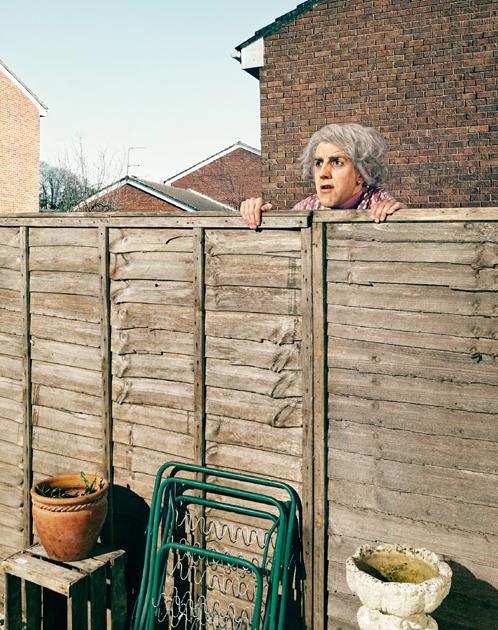 An elderly woman with grey hair and a pink cardigan peering over a wooden fence, looking surprised.