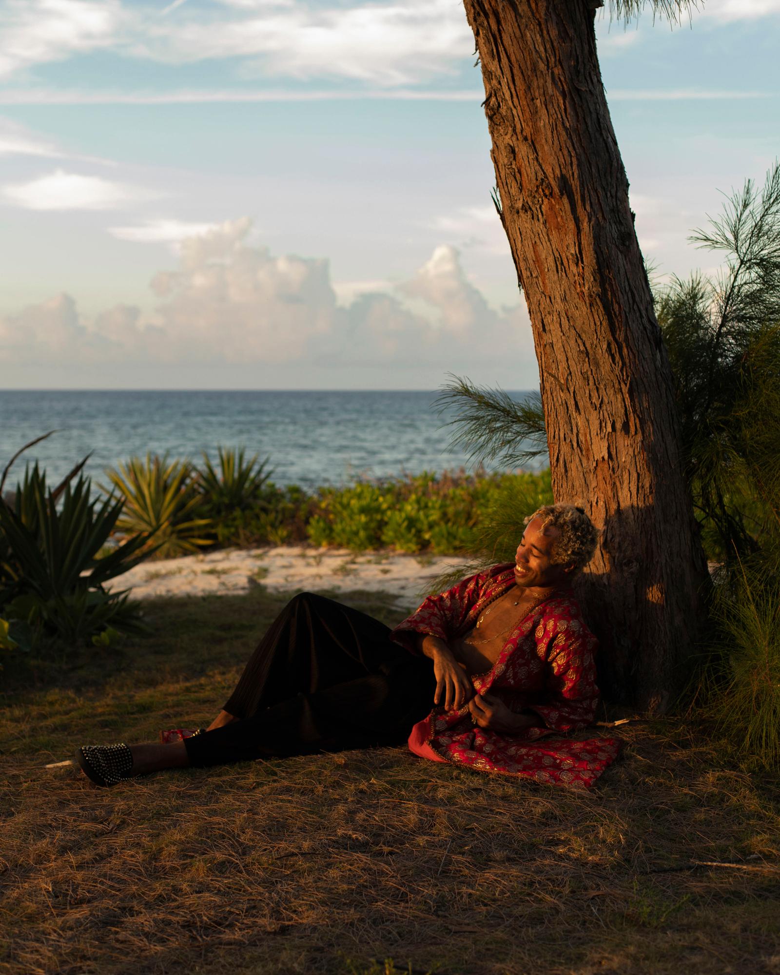A person is lying on the ground, surrounded by lush greenery and a body of water in the background.