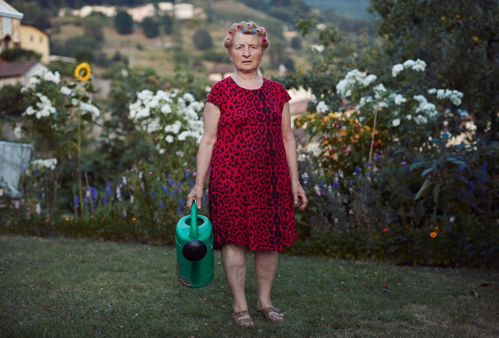 An elderly woman in a red dress with leopard print, holding a watering can, standing in a garden with flowers and a house in the background.