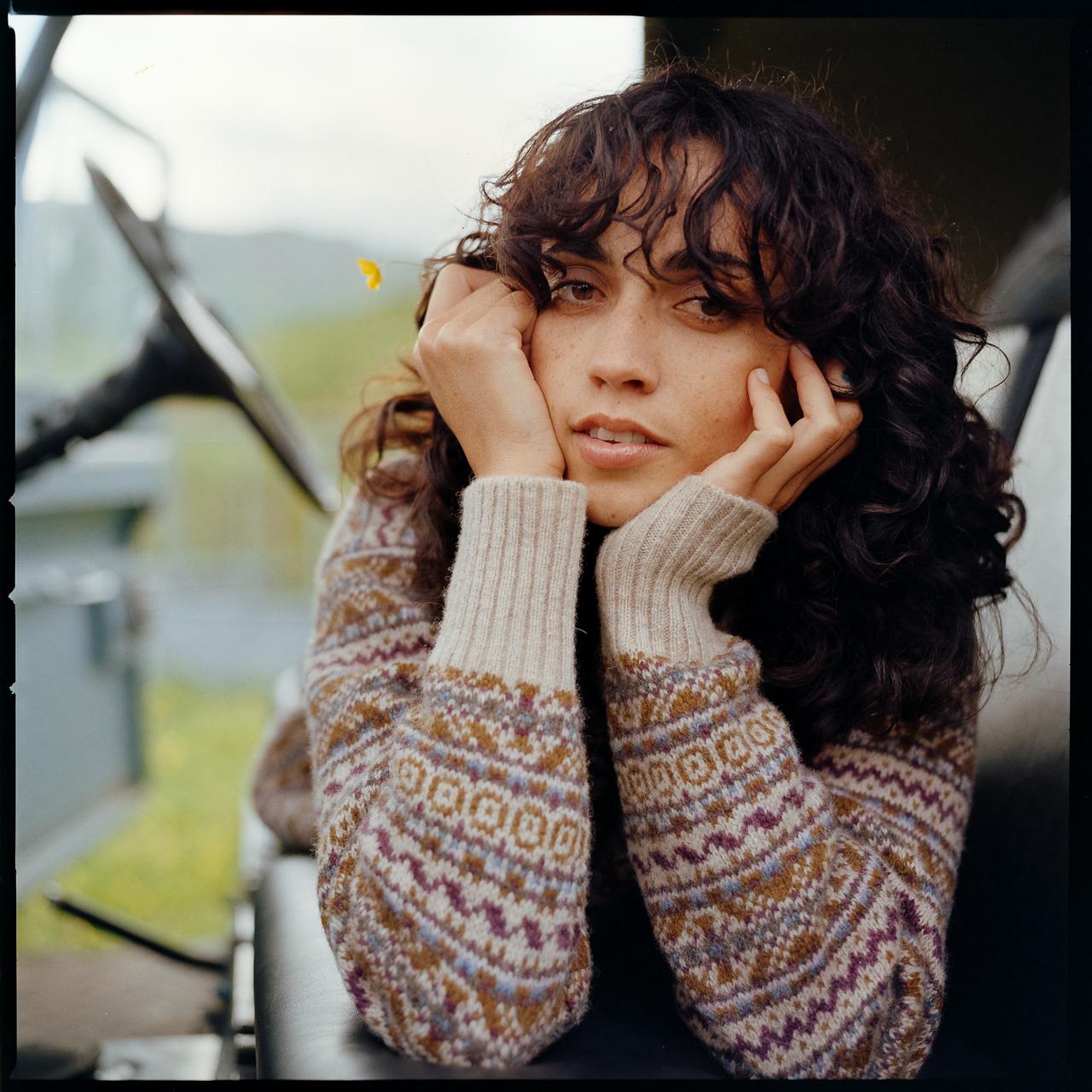 A woman with curly brown hair and a patterned sweater, sitting in a chair with her hands on her face, looking out of a window with a blurred landscape in the background.