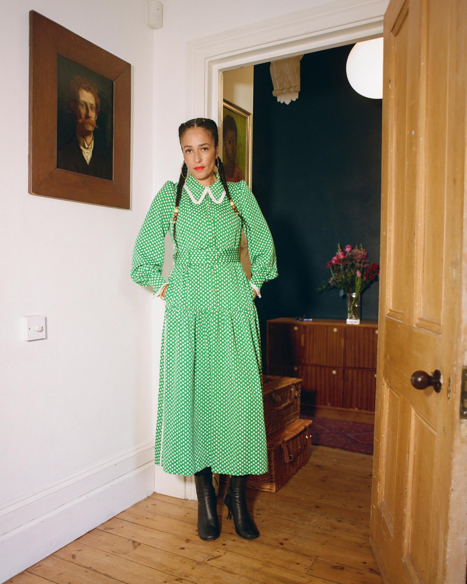 A woman with dark hair in a green dress with white polka dots, standing in a room with a white wall and a portrait on the wall.