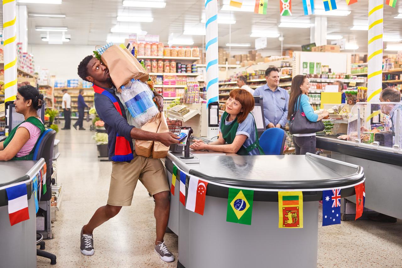 A man in a blue shirt and khaki shorts is carrying multiple shopping bags from a supermarket checkout.