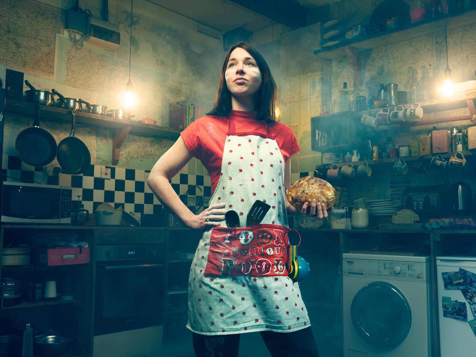 A woman in a kitchen, wearing a red and white polka dot apron, holding a freshly baked loaf of bread.